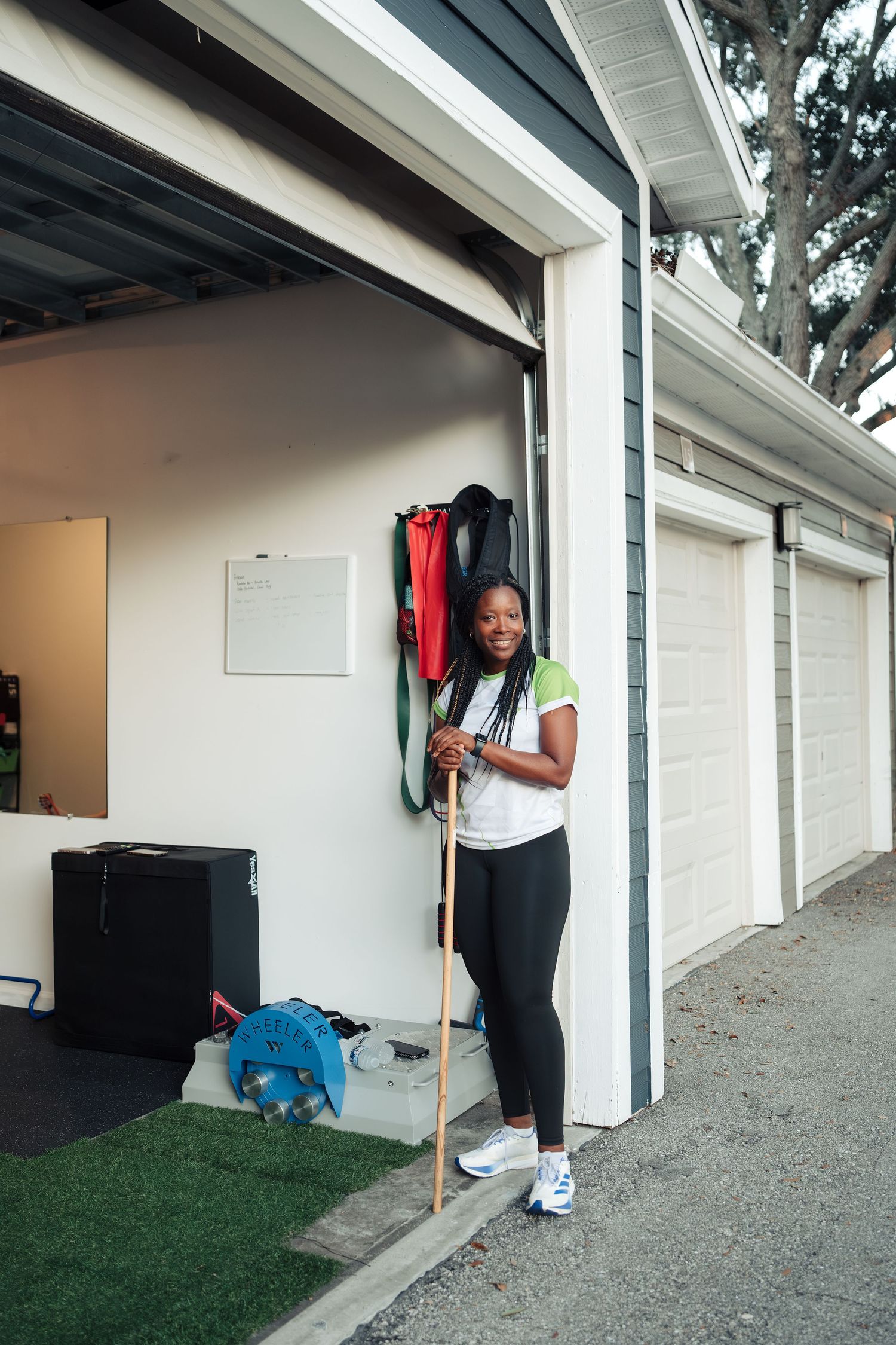 Woman in athletic wear holding a wooden staff outside a home garage gym with resistance bands and equipment.