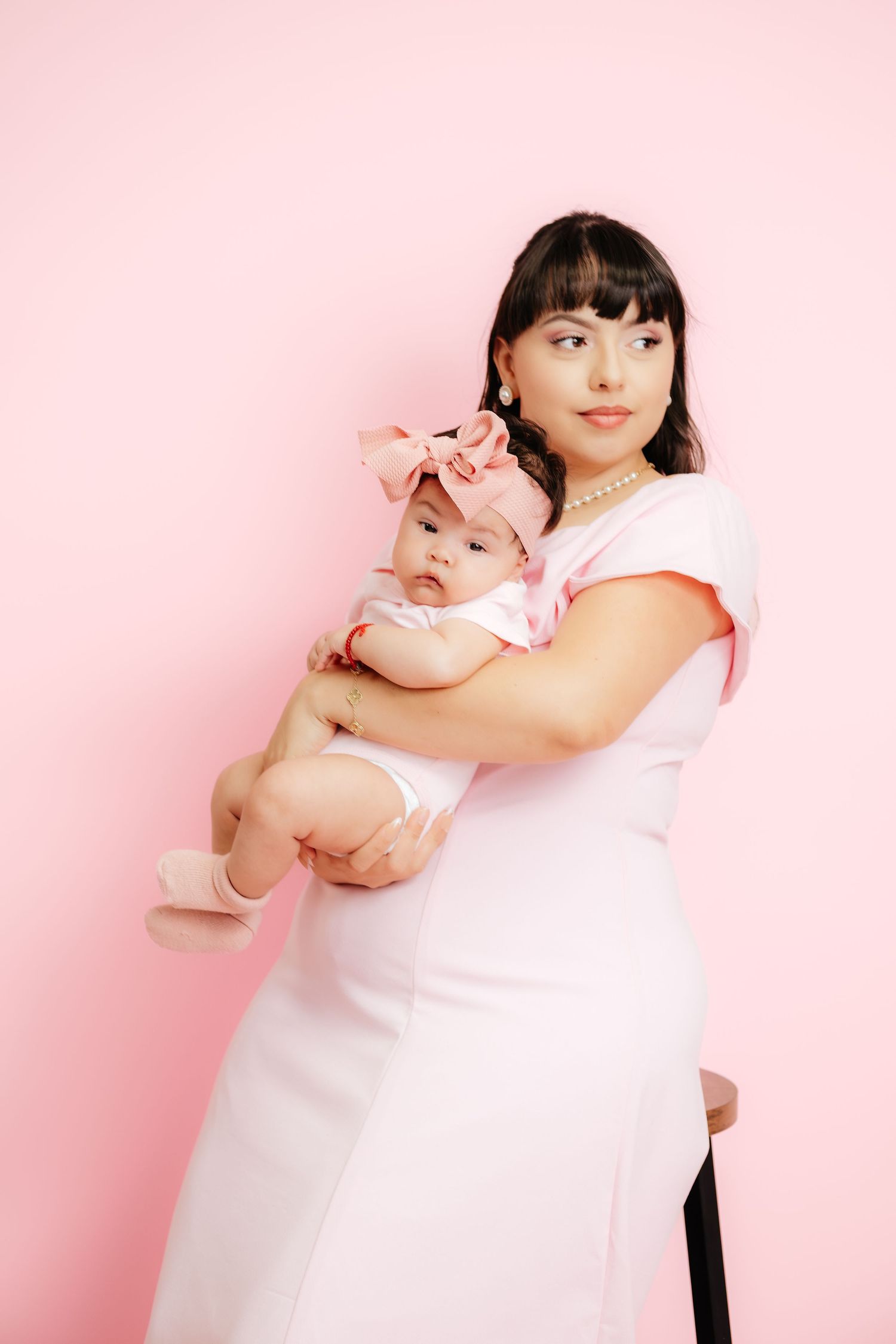 A young mother in a pink dress holds her baby girl with a pink bow headband against a pink background.