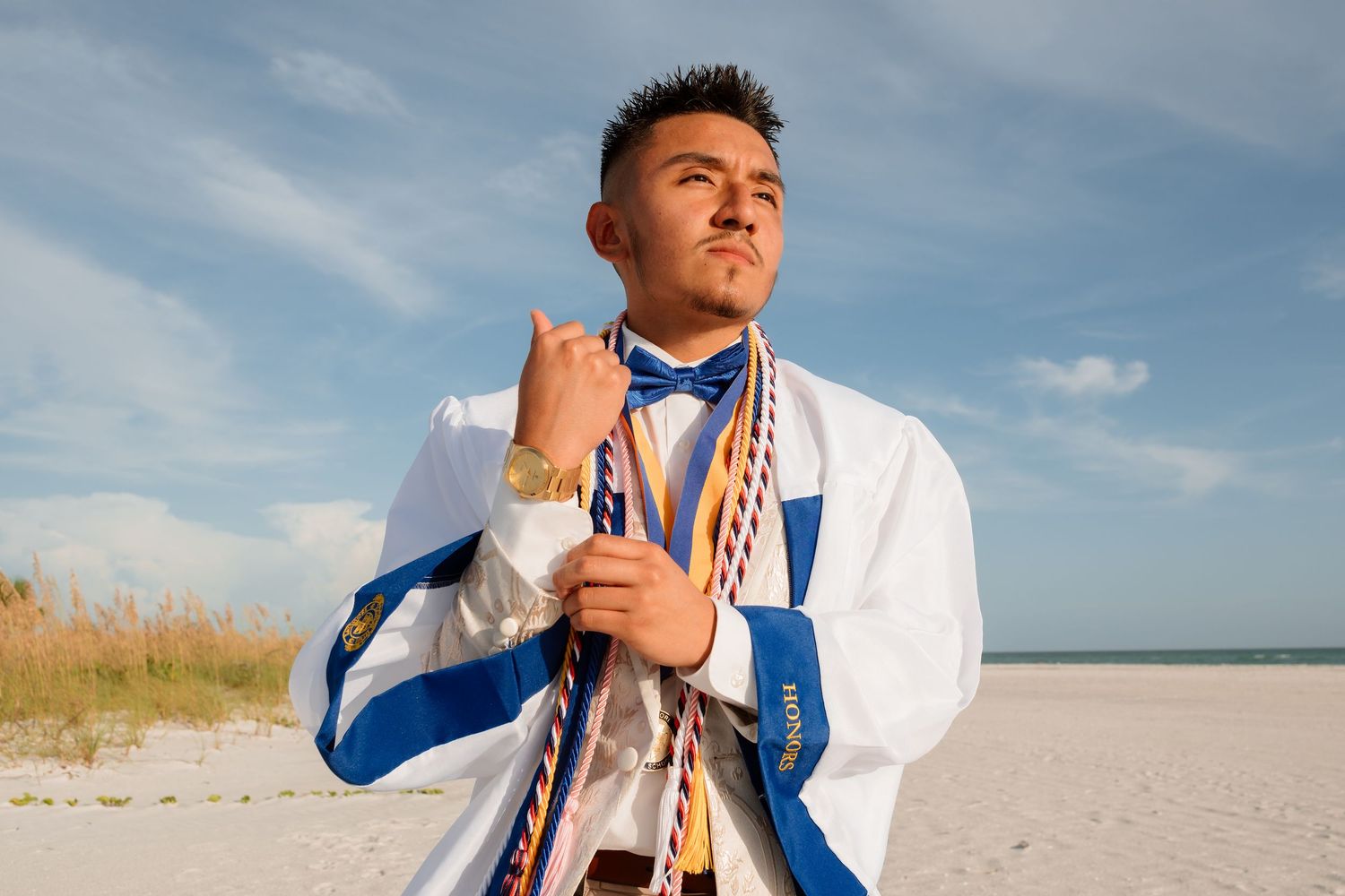 Young man in white and blue graduation attire with medals poses confidently on a sunny beach.
