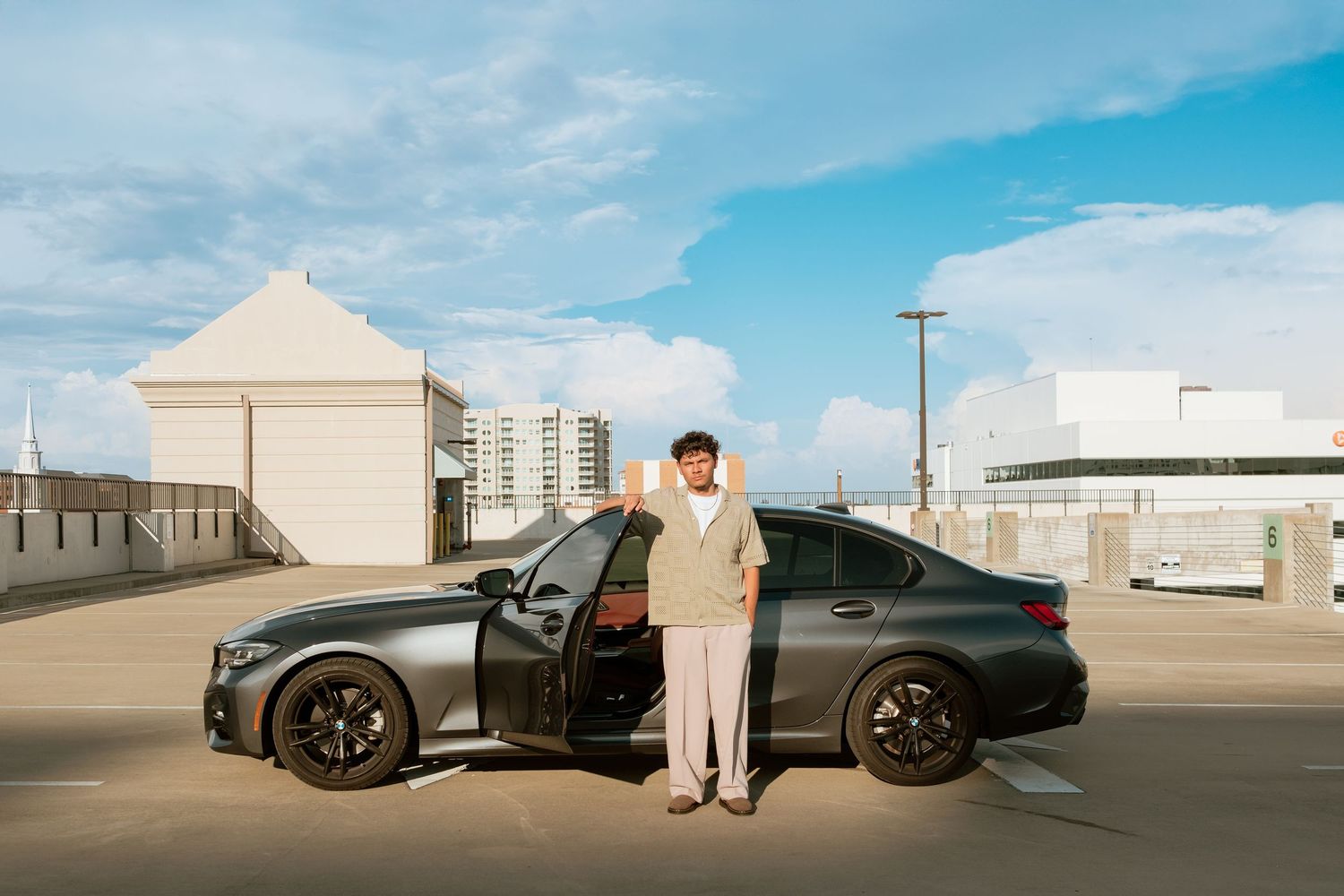 Man in beige suit standing beside a dark gray BMW sedan in an open parking lot under a blue sky.