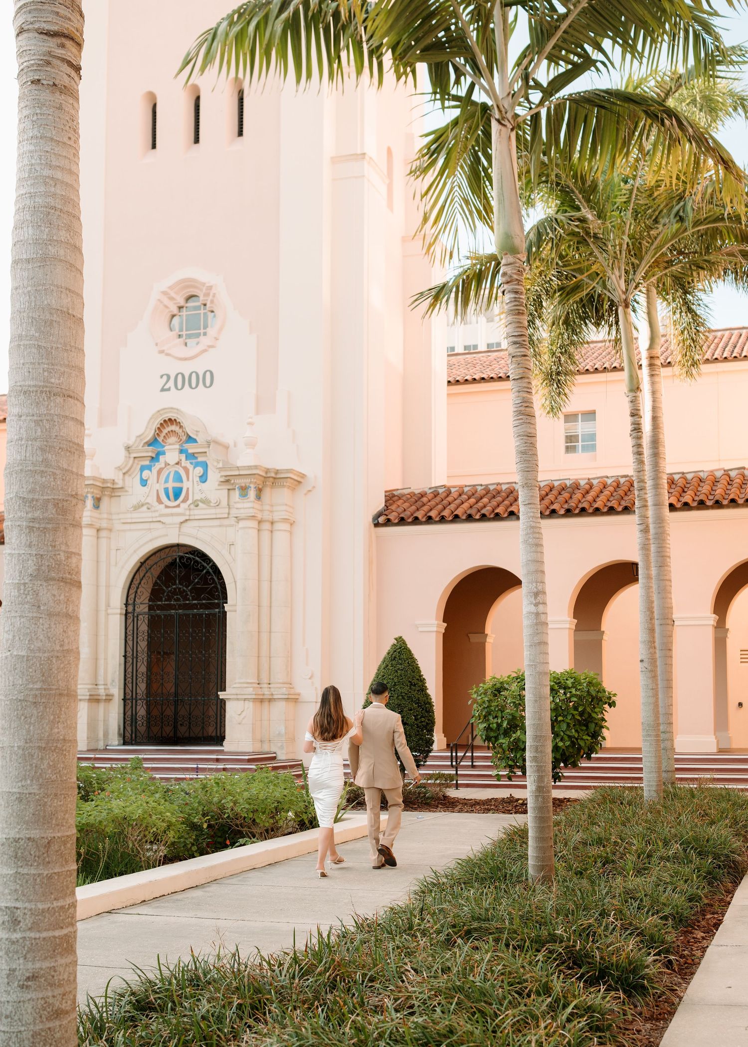 Couple walks hand-in-hand toward a pink Spanish-style building with palm trees and arched colonnades.
