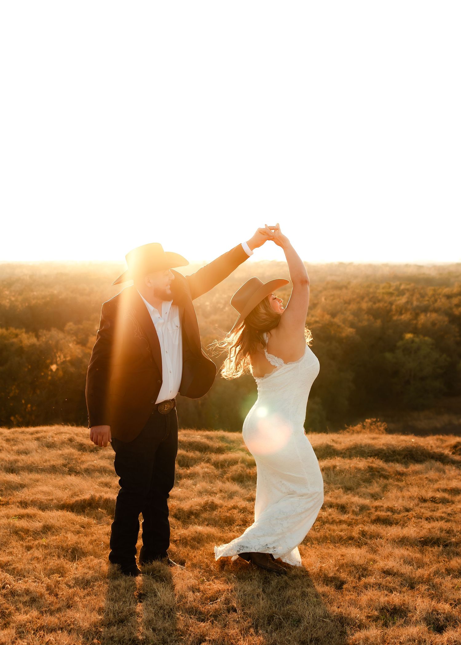 Couple dancing at golden hour sunset in a field, bride in white gown and groom in black suit with cowboy hat.