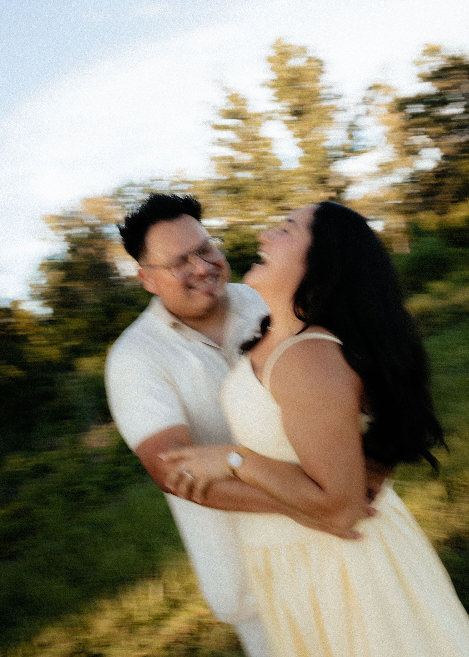 A joyful couple embraces and laughs outdoors with a motion blur effect, surrounded by lush green trees.