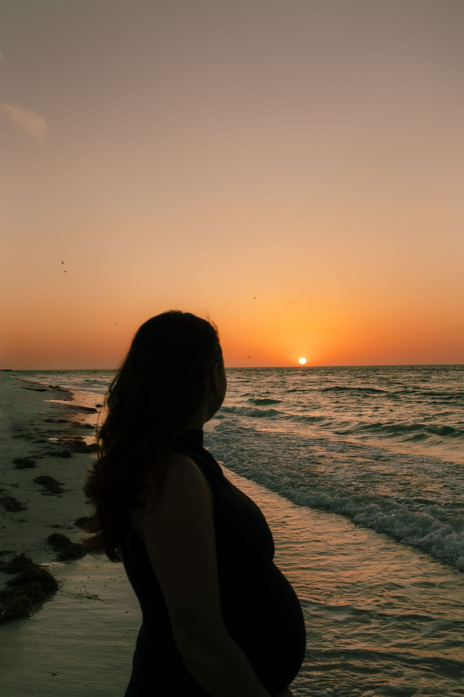 Silhouetted figures embrace on a beach at sunset as waves crash along the shoreline against an orange sky.