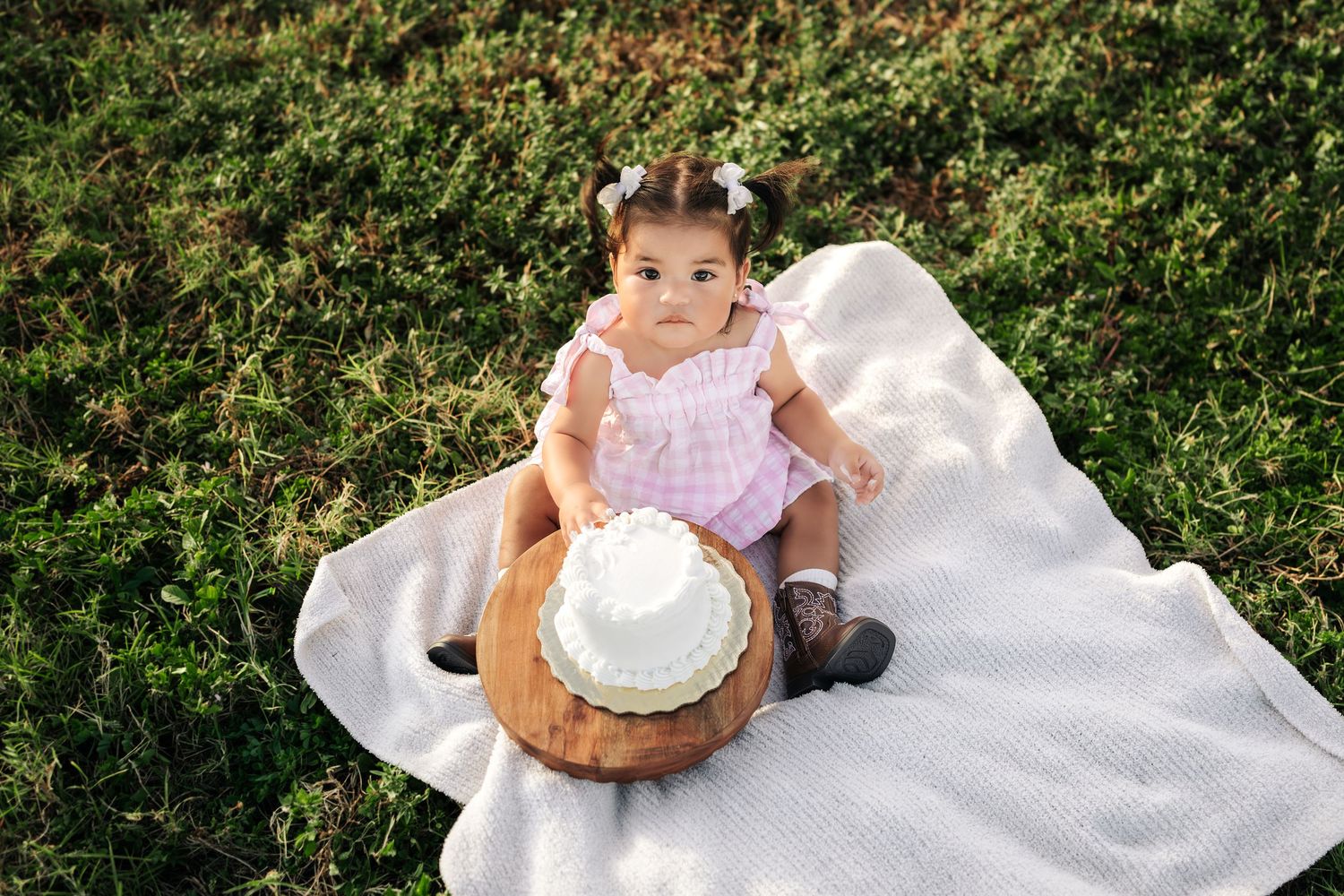 Baby in a white dress sits on grass with a cake during a first birthday photo session in a garden setting.