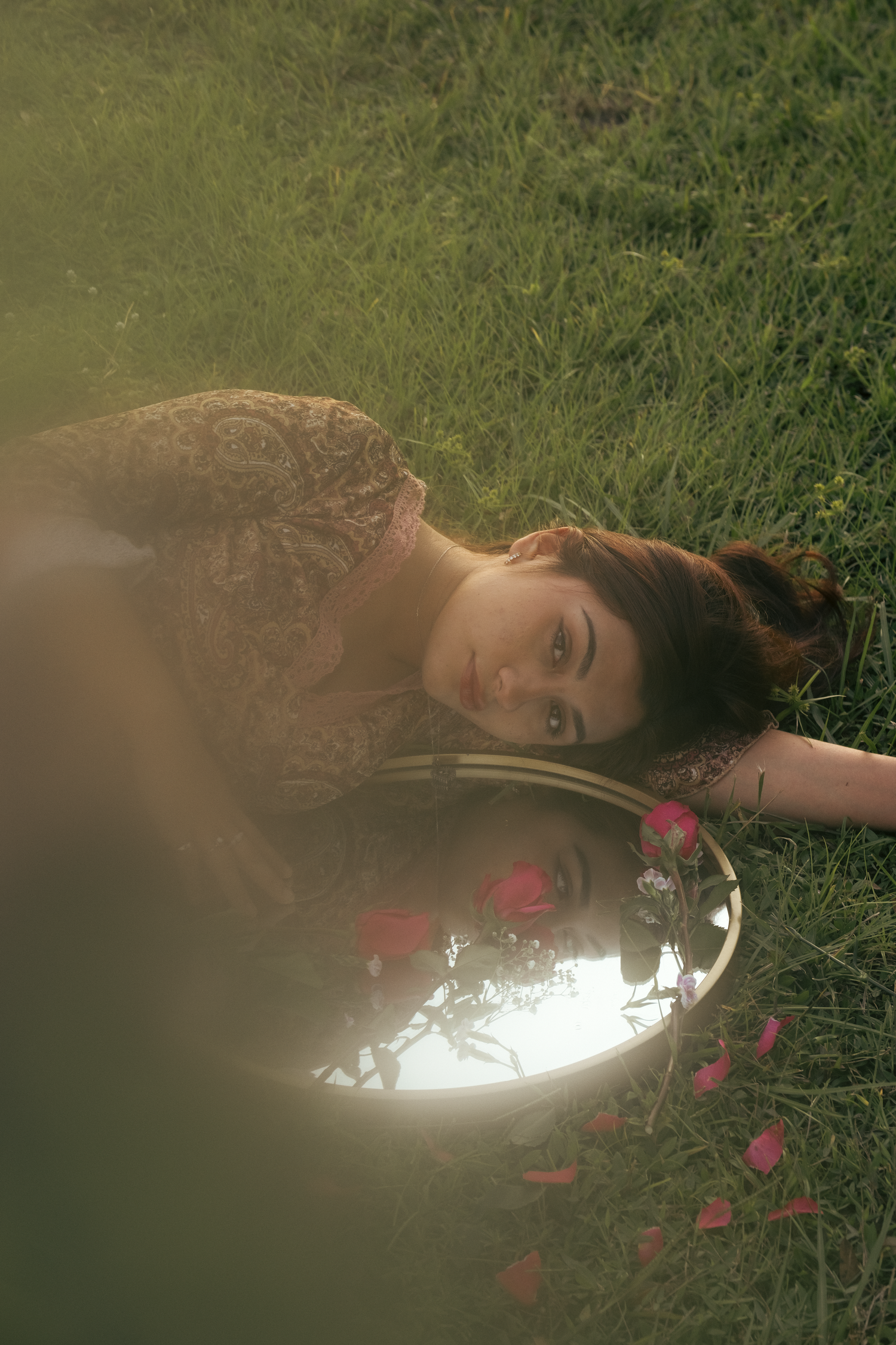 Dreamy portrait lying beside reflective mirror with flowers in soft golden light.