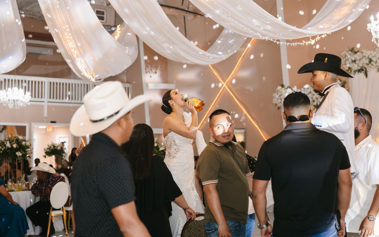 Couple sharing a drink at elegant wedding reception with white draping, guests in cowboy hats celebrating around them.