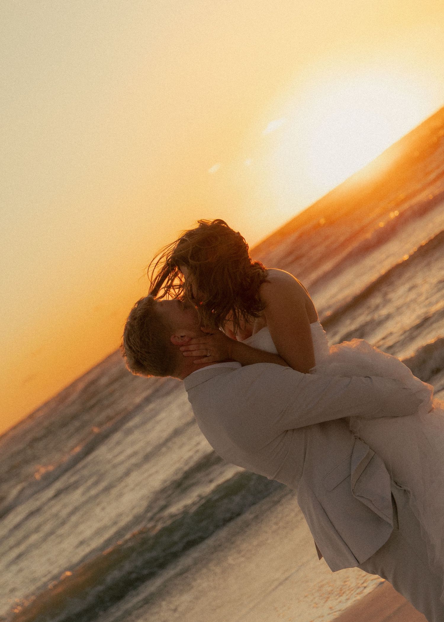 Couple sharing a romantic kiss on the beach during a golden sunset, waves gently crashing in the background.