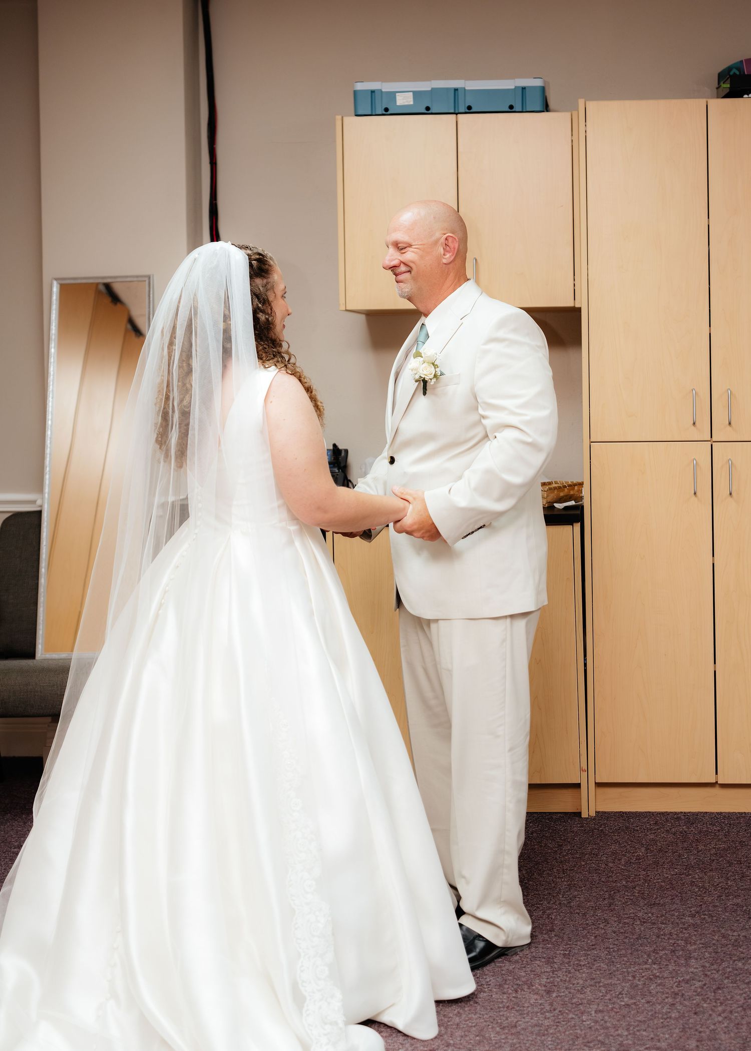 Bride in white gown and veil holds hands with bald groom in cream suit during first look moment.
