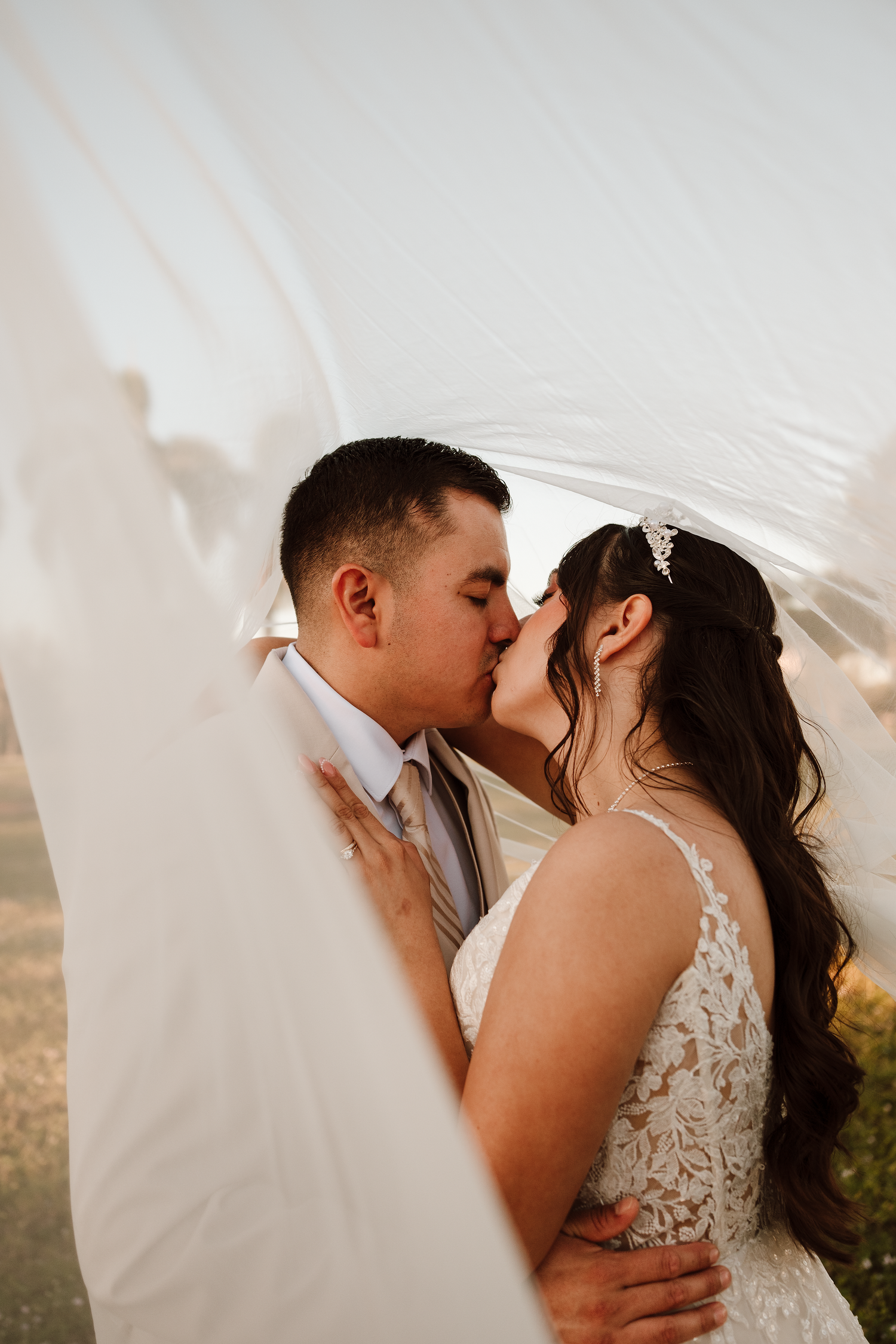 Bride and groom share a romantic kiss under a flowing white veil during their outdoor wedding ceremony.