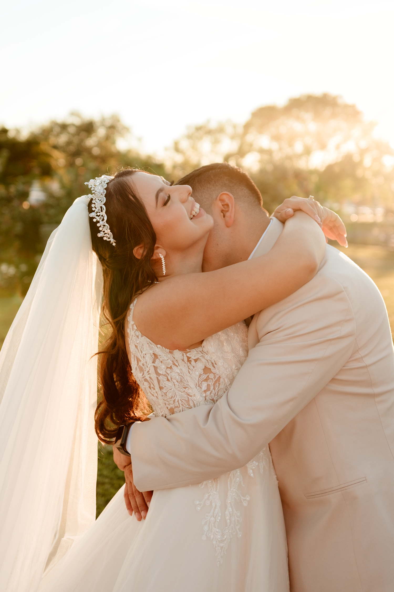 Bride in lace gown with tiara embracing groom in warm golden sunset light at outdoor wedding.