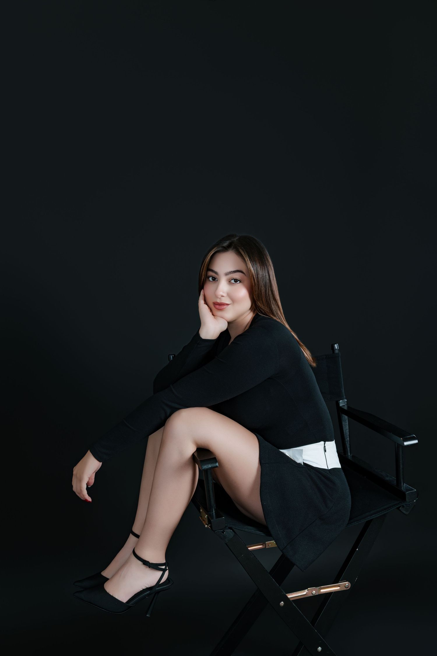 Young woman in black outfit sitting gracefully on a director's chair against a dark background.