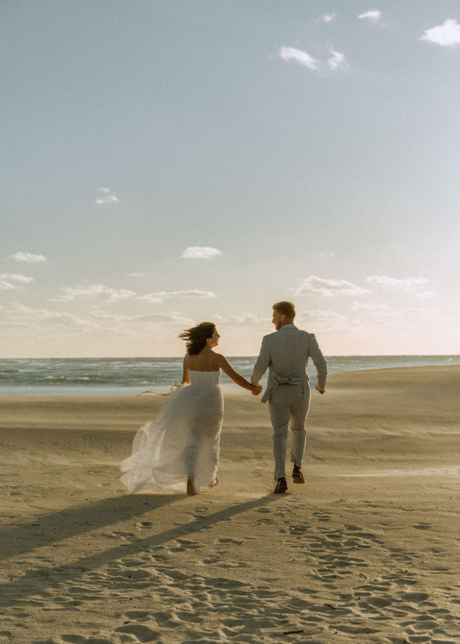 Bride and groom holding hands running on sandy beach at sunset in wedding attire.