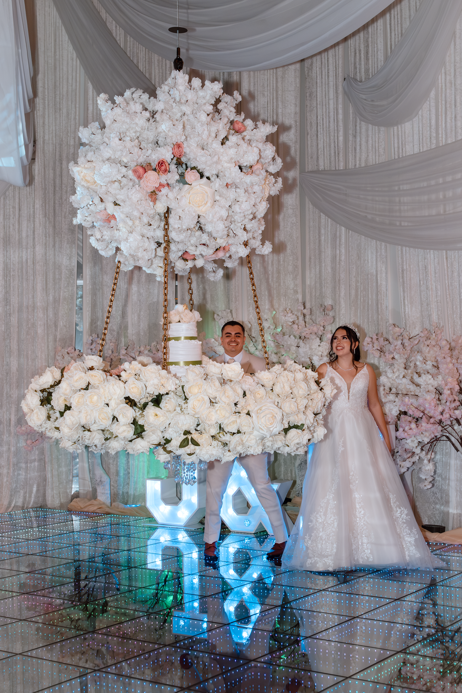Bride and groom posing beside suspended floral wedding cake display with illuminated initials on mirrored dance floor.