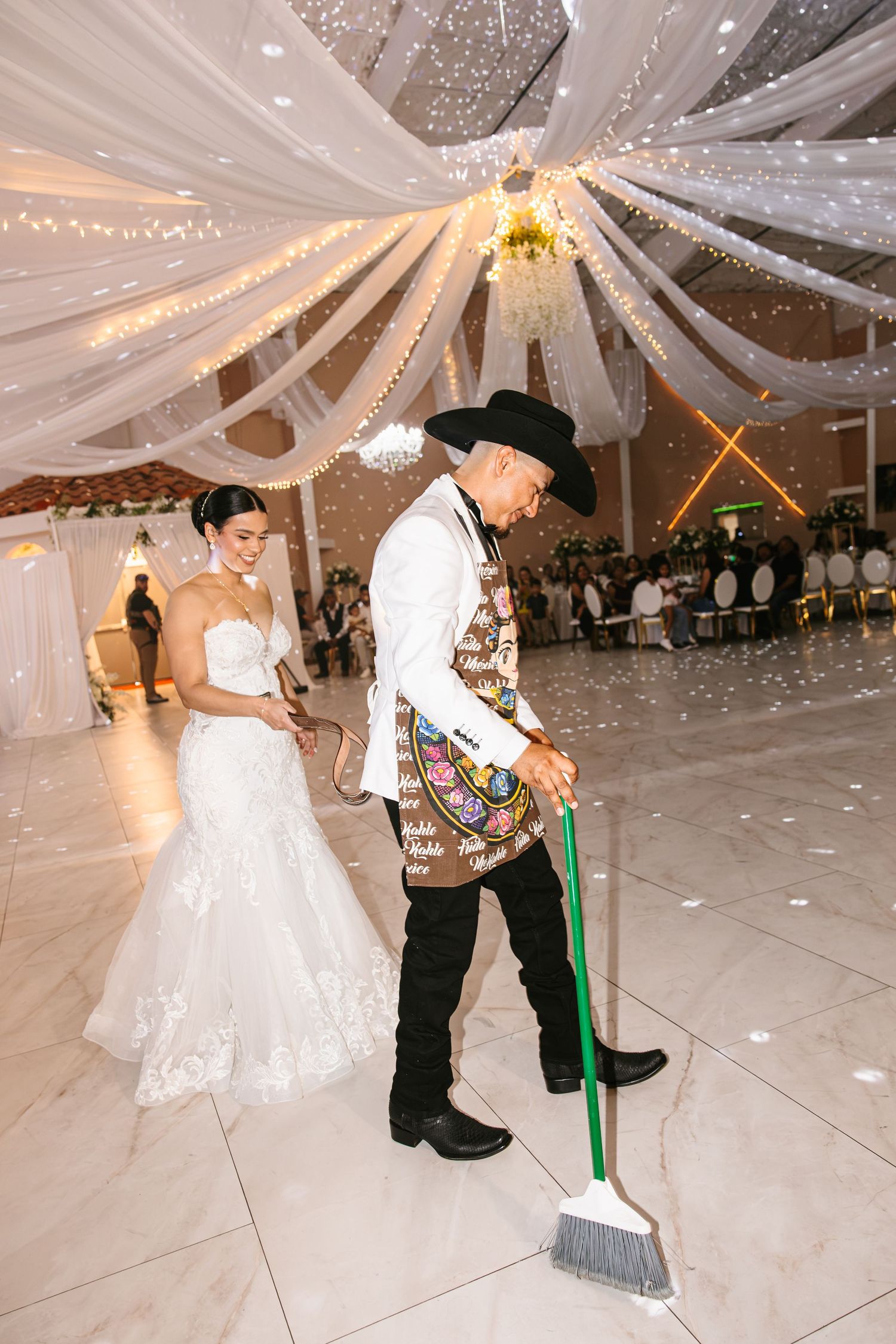 Groom in cowboy hat sweeping dance floor as bride watches at elegant wedding reception.