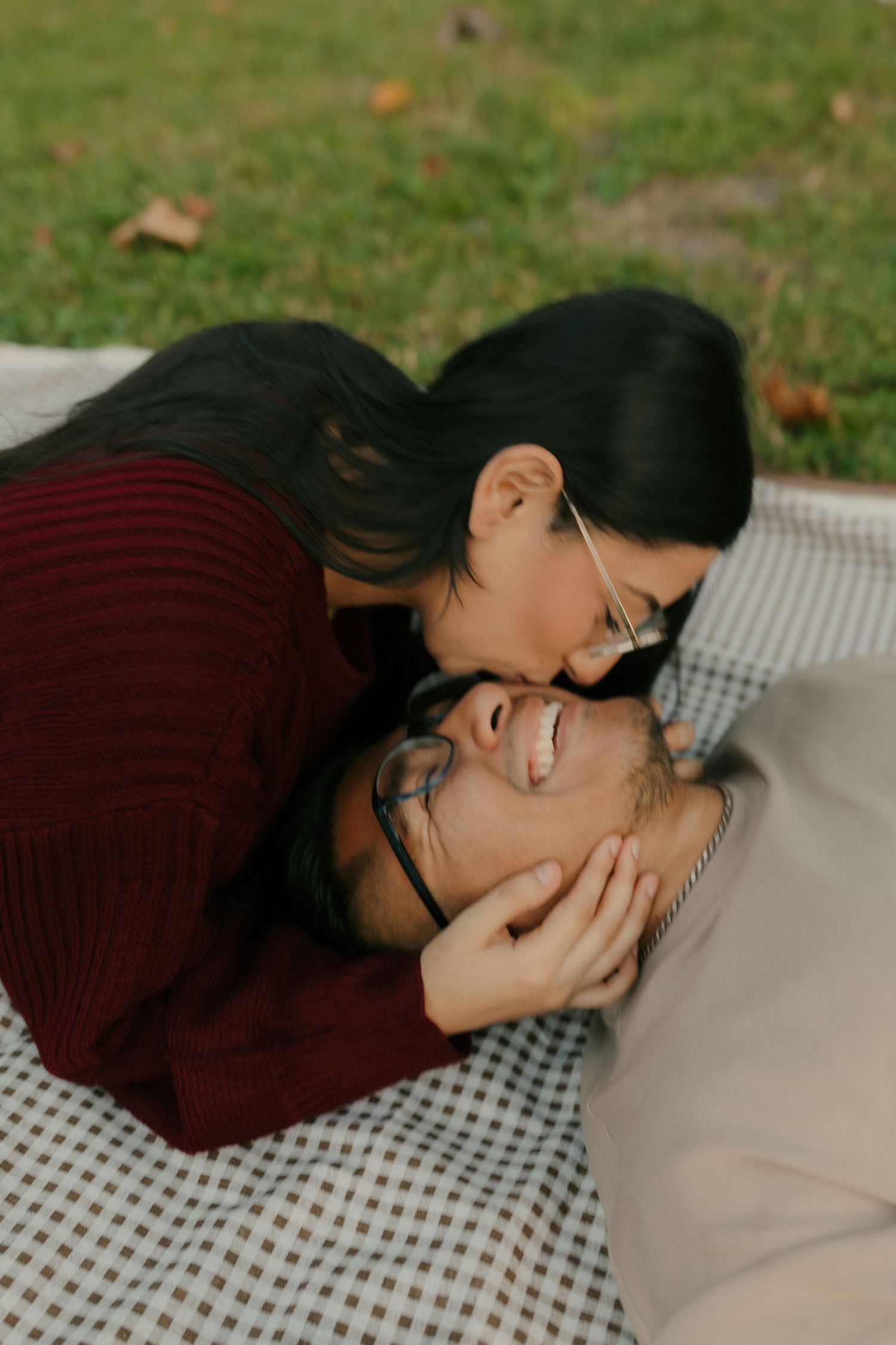 Smiling man with glasses lies on a checkered picnic blanket as a woman in a burgundy sweater leans down to kiss his forehead on the grass.