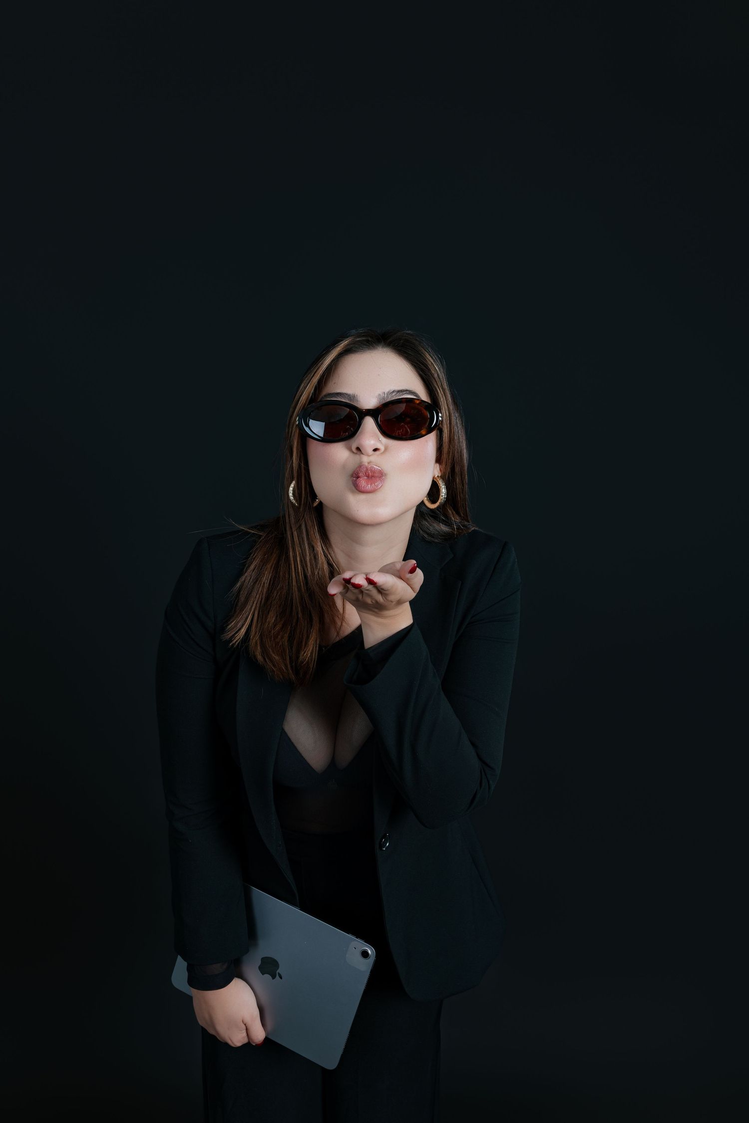 Stylish woman in black blazer and sunglasses blowing a kiss while holding a laptop against dark background.