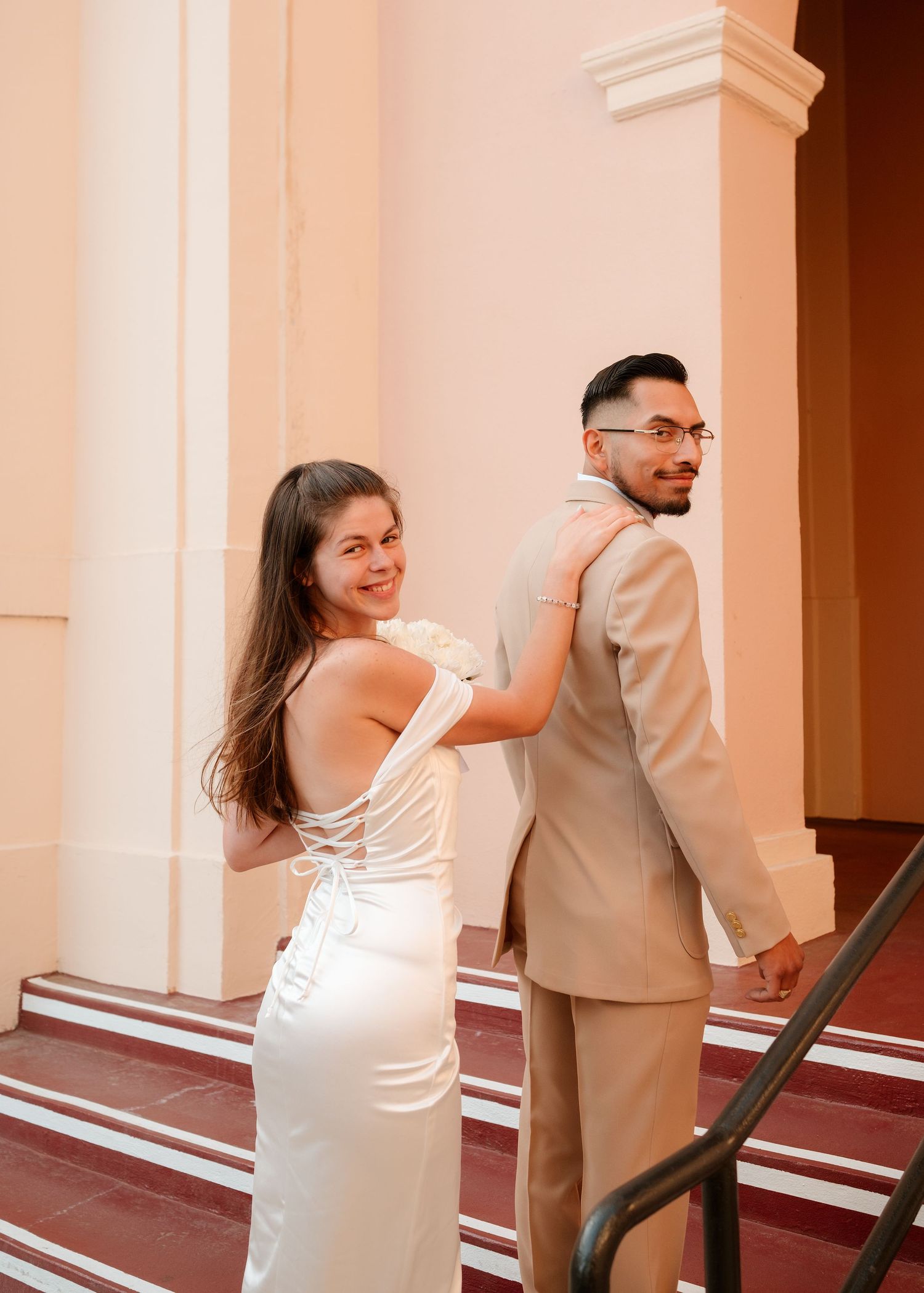 Couple in wedding attire poses on steps, woman in white gown smiles back, man in beige suit stands forward.