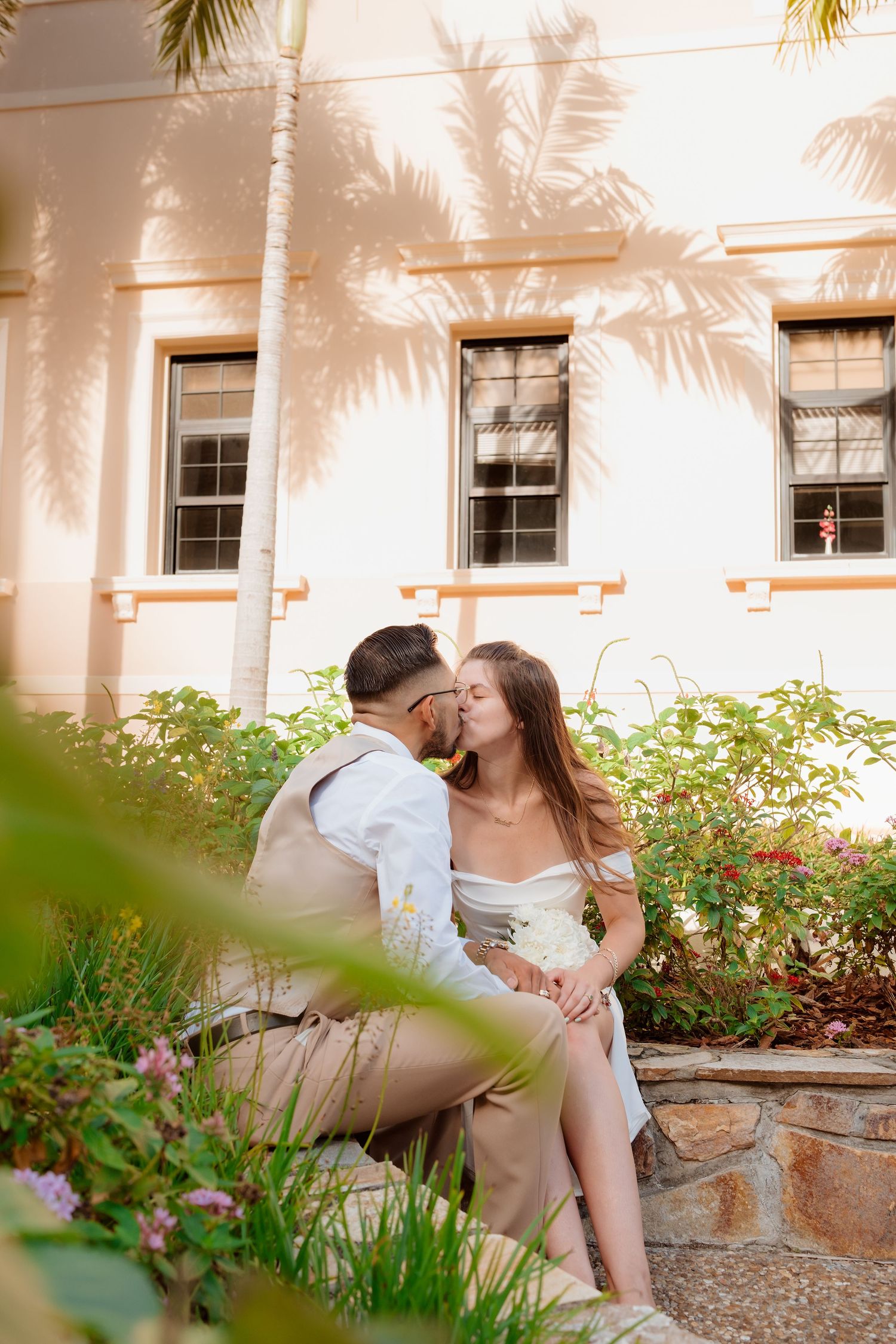 Romantic couple shares a tender kiss while seated in a lush garden beside a pink building.