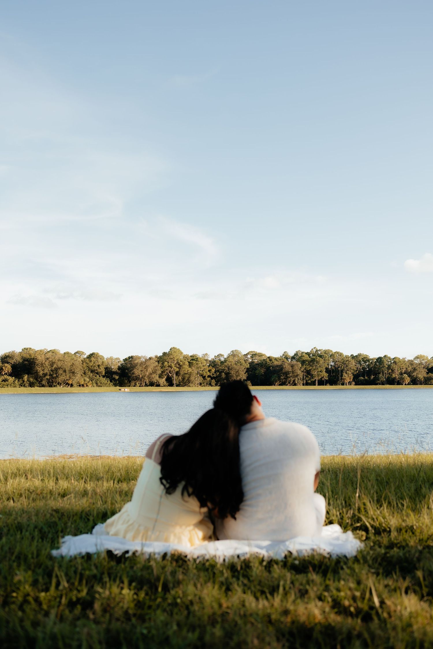 Couple sitting together on a blanket by a serene lake, surrounded by lush greenery under a clear blue sky.