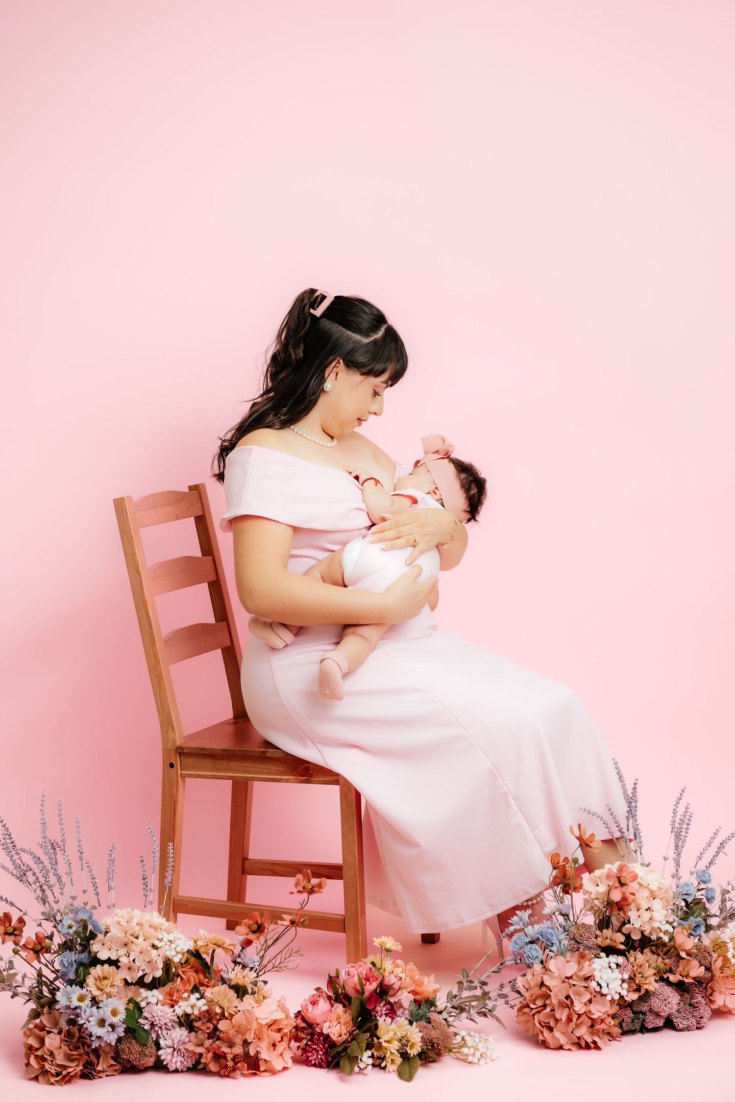 A mother in a white dress sits on a wooden chair cradling her newborn surrounded by colorful flowers on pink.