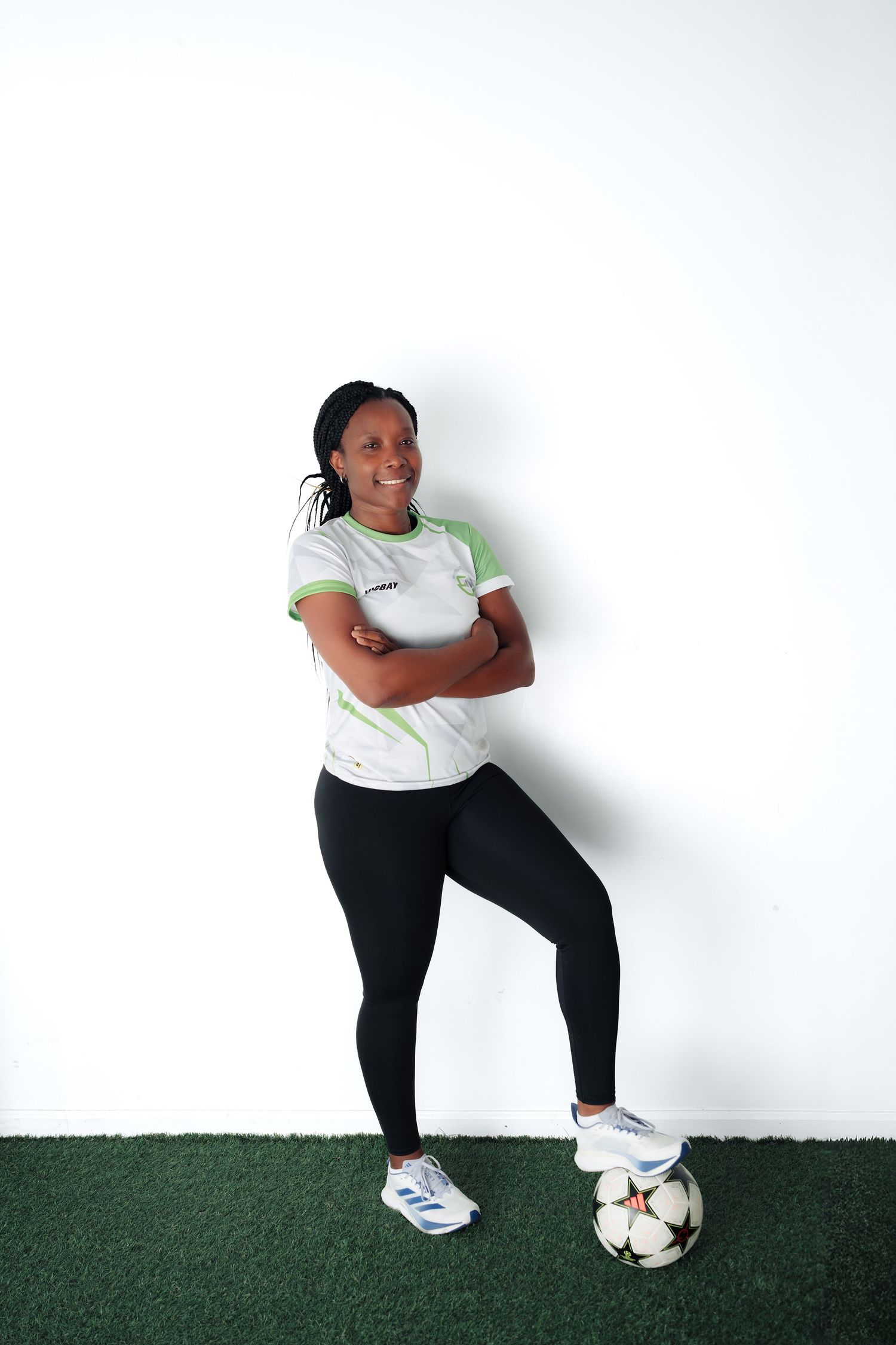 Female soccer player in white and green jersey poses confidently with arms crossed, foot resting on a soccer ball.