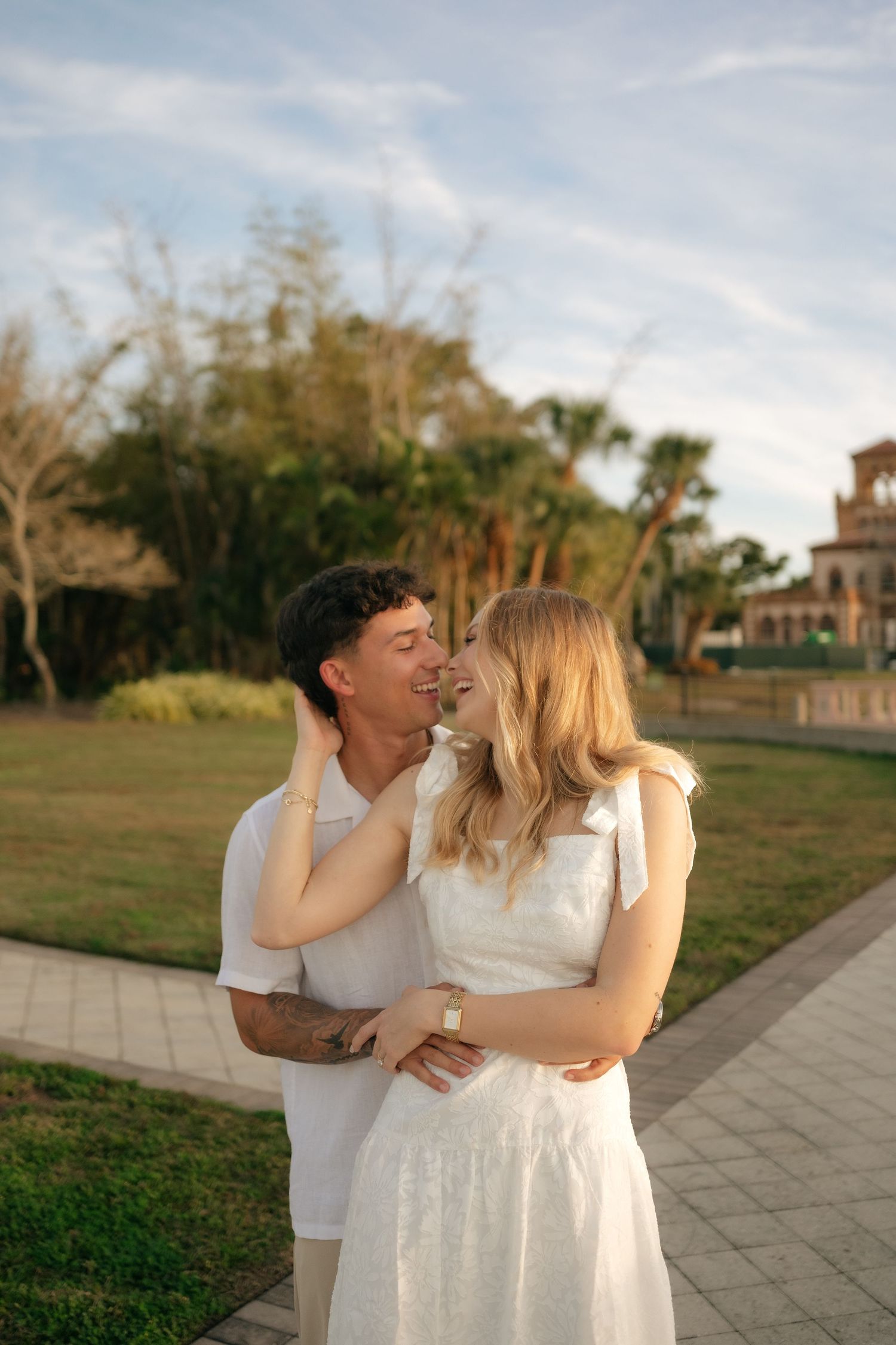 A couple in white outfits share a romantic moment outdoors at golden hour, surrounded by lush greenery.