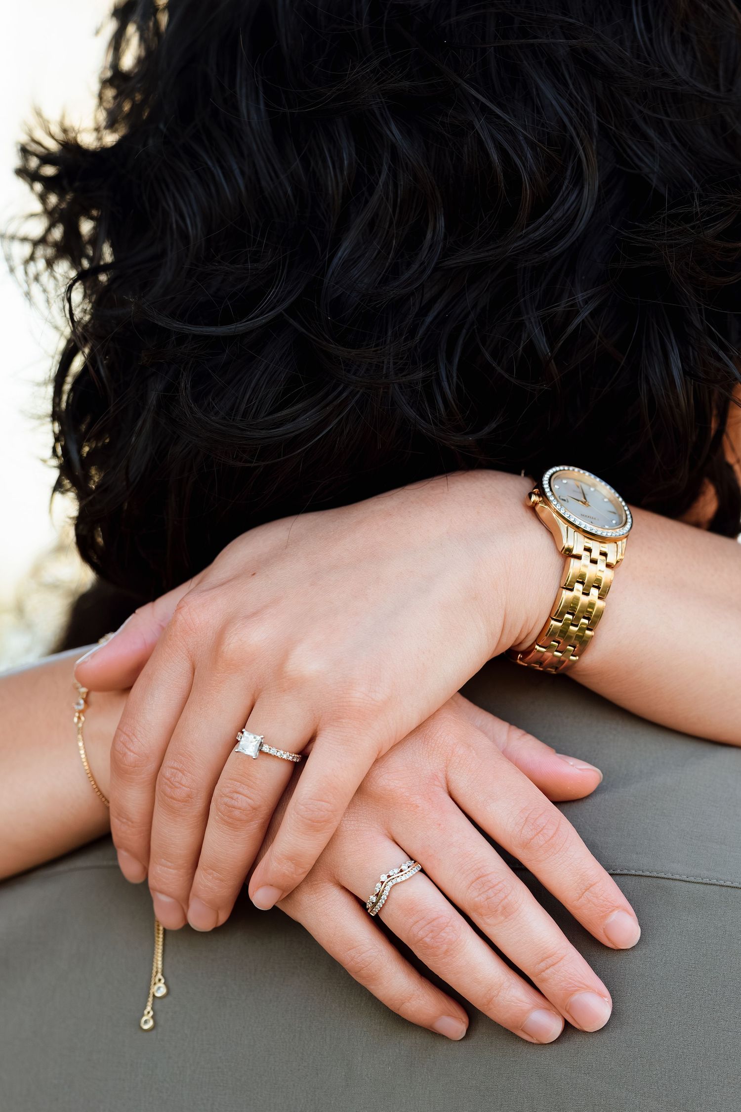 Two hands adorned with diamond rings, a gold chain bracelet, and a gold watch rest on a persons shoulder.
