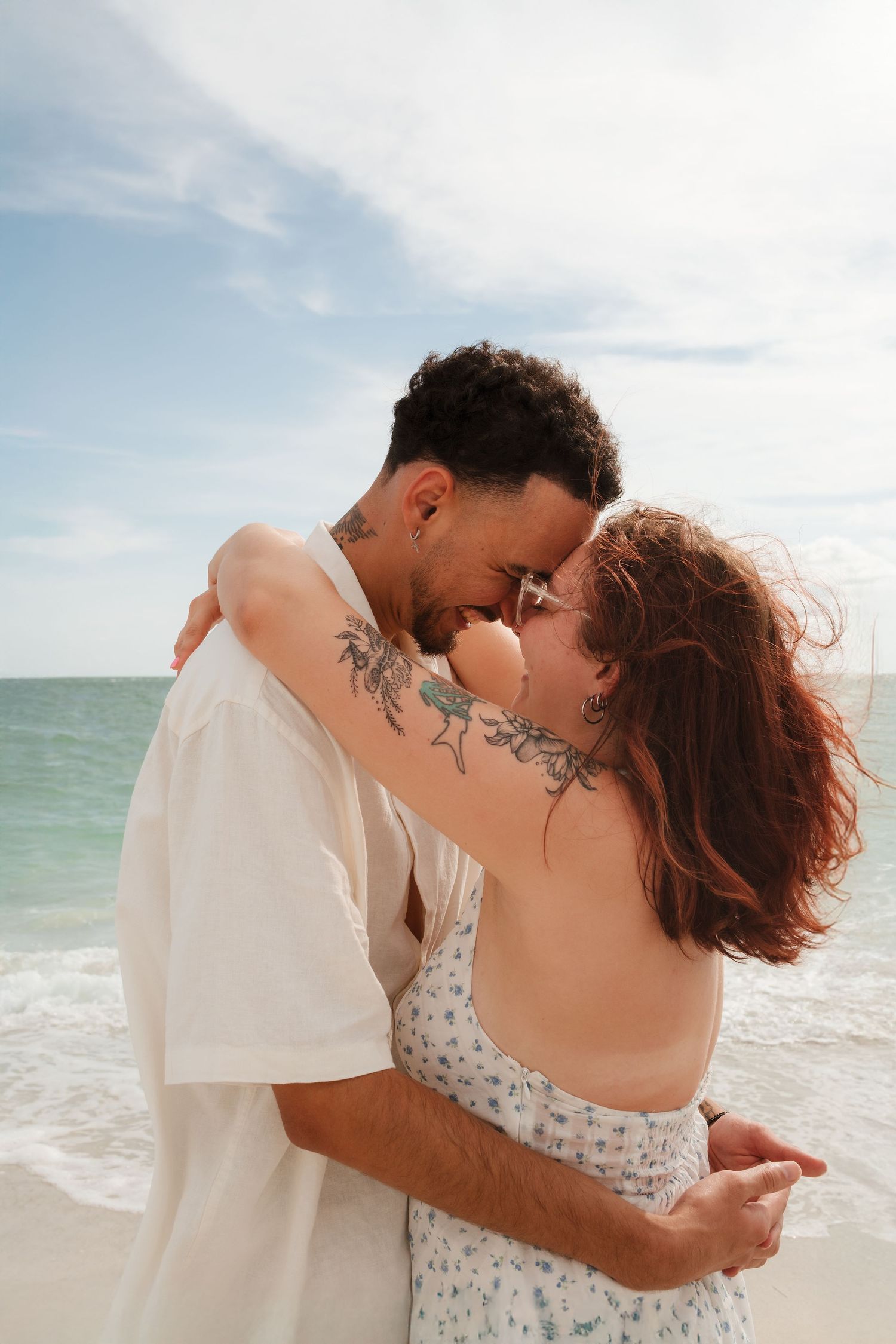 Couple embracing and smiling on a sunny beach, man with tattoos in white shirt, woman in floral dress with red hair.