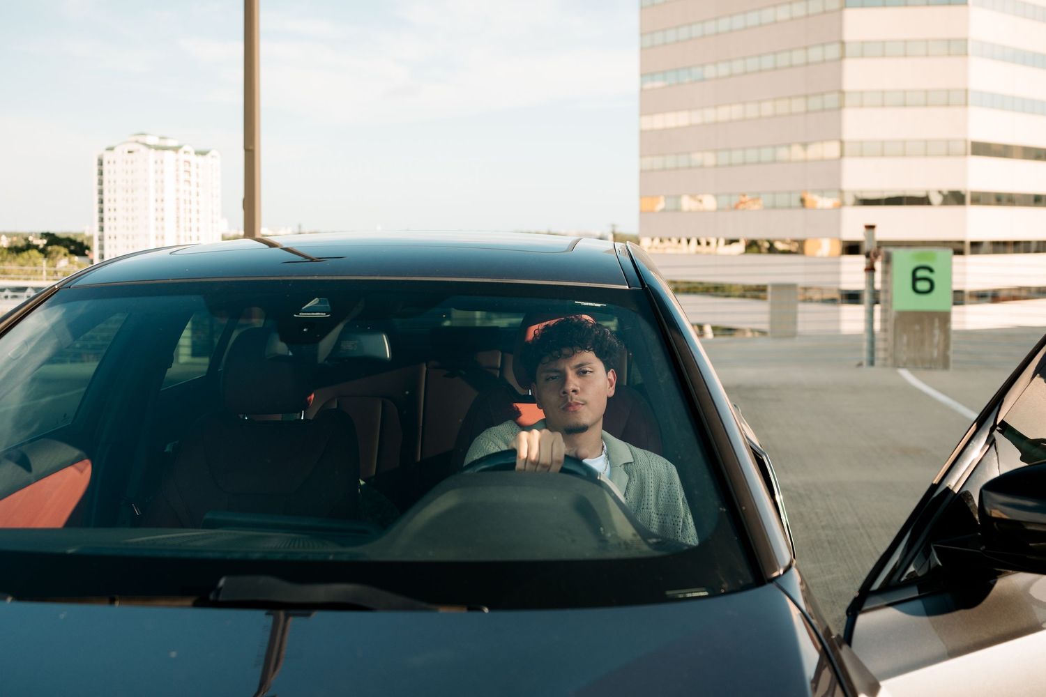 Man sitting in driver's seat of black car in a parking garage with city buildings in background.