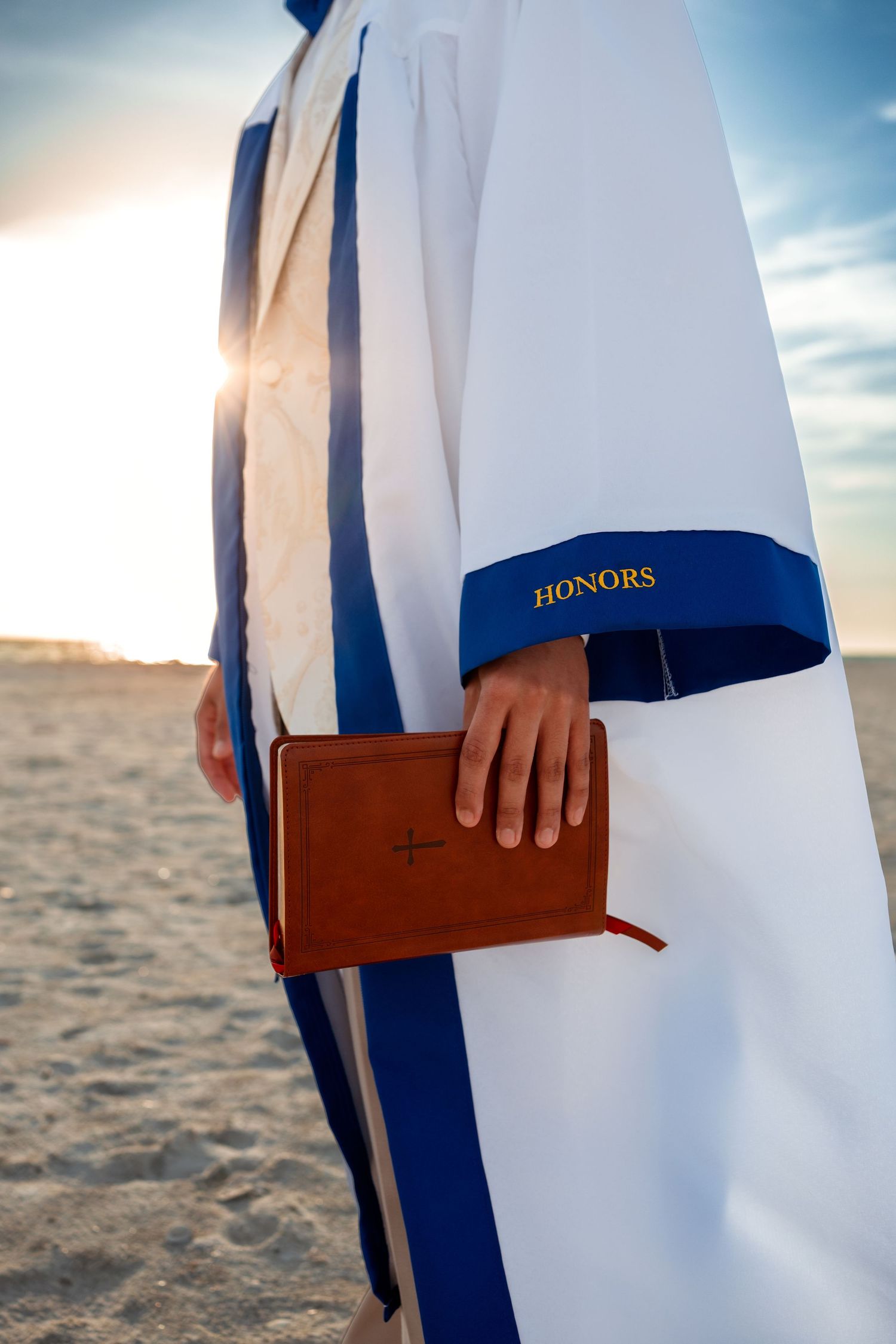 Graduate in honors regalia holding a Bible on a sandy beach at sunset.