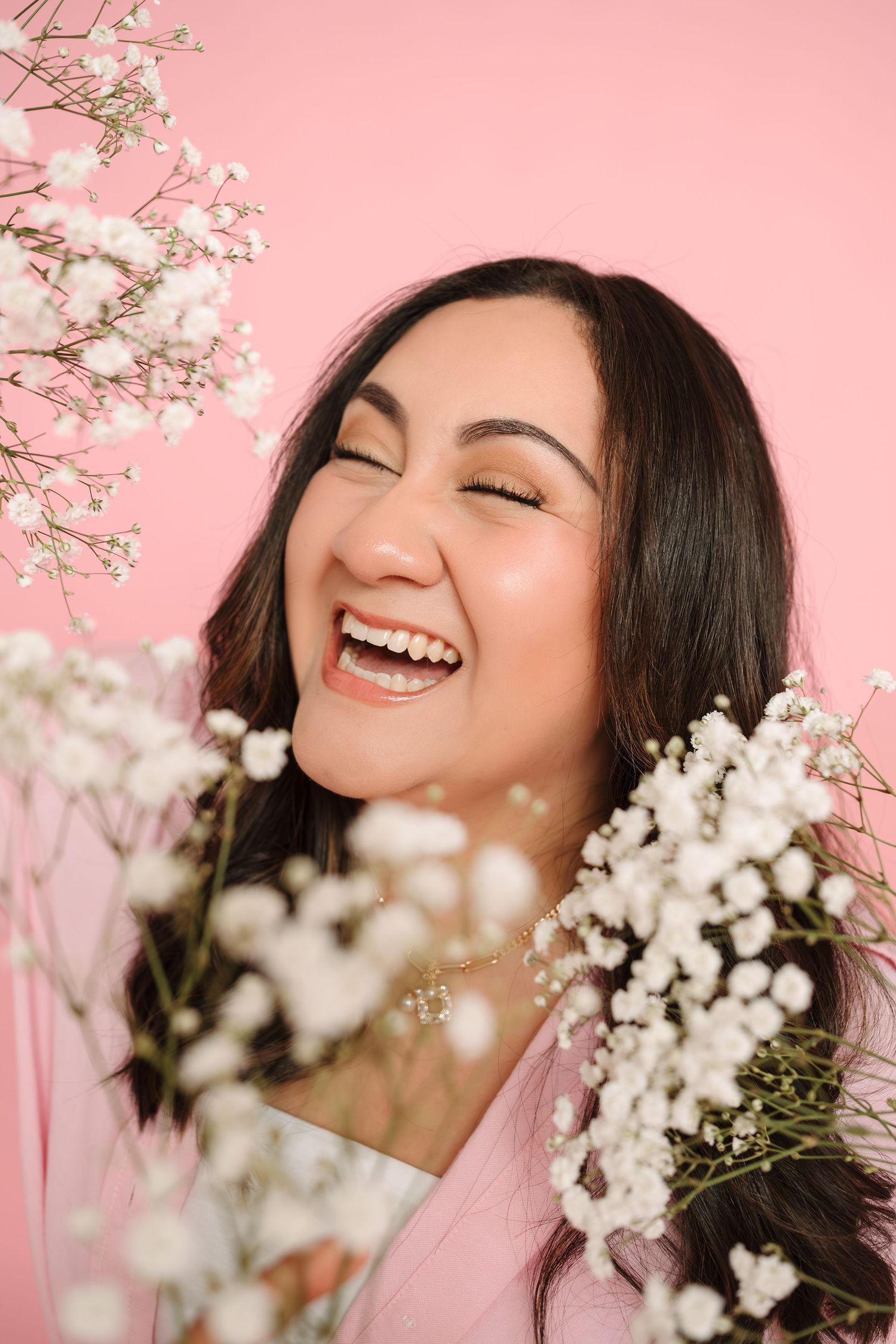 A joyful Asian woman laughing with eyes closed, surrounded by white baby's breath flowers against a pink background.