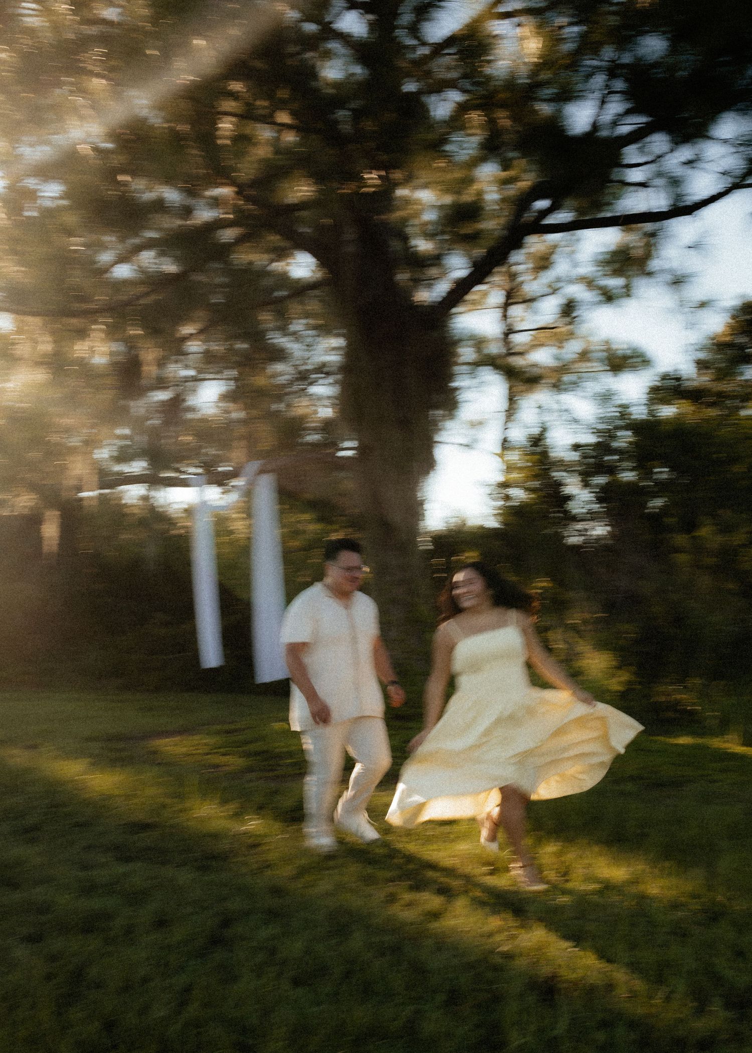 Couple in white attire running joyfully through sunlit garden with large tree backdrop at golden hour.