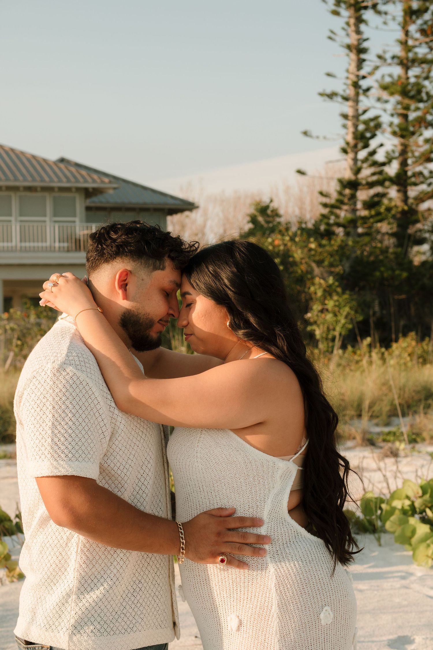 A couple in matching white outfits embrace tenderly on a sandy beach at golden hour with trees and a house behind them.