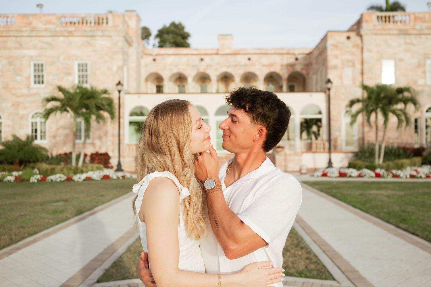 Couple sharing a romantic moment in front of an elegant estate with palm trees and manicured gardens.