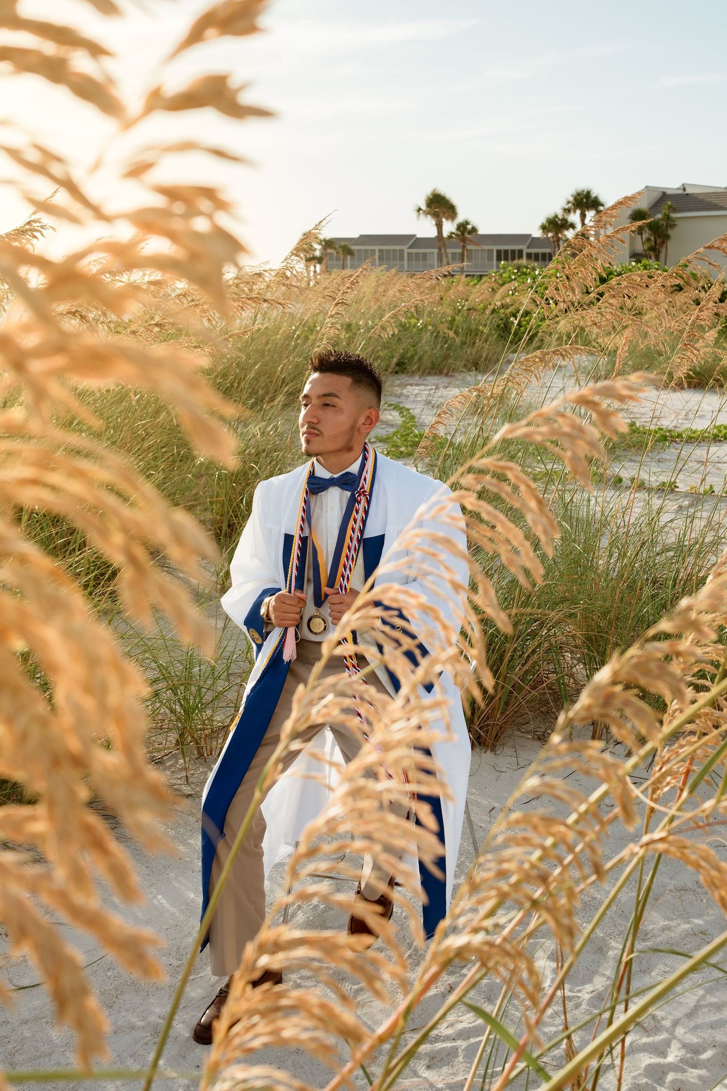 Young man in white graduation gown with blue sash and medals stands among golden sea oats on a sandy beach at sunset.