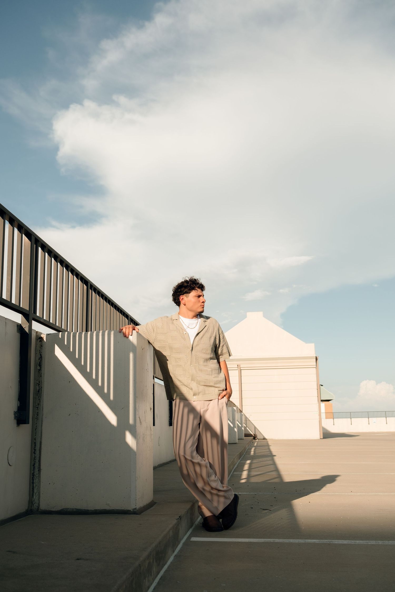 A young man in a striped suit leans against a railing on a rooftop under a partly cloudy blue sky.