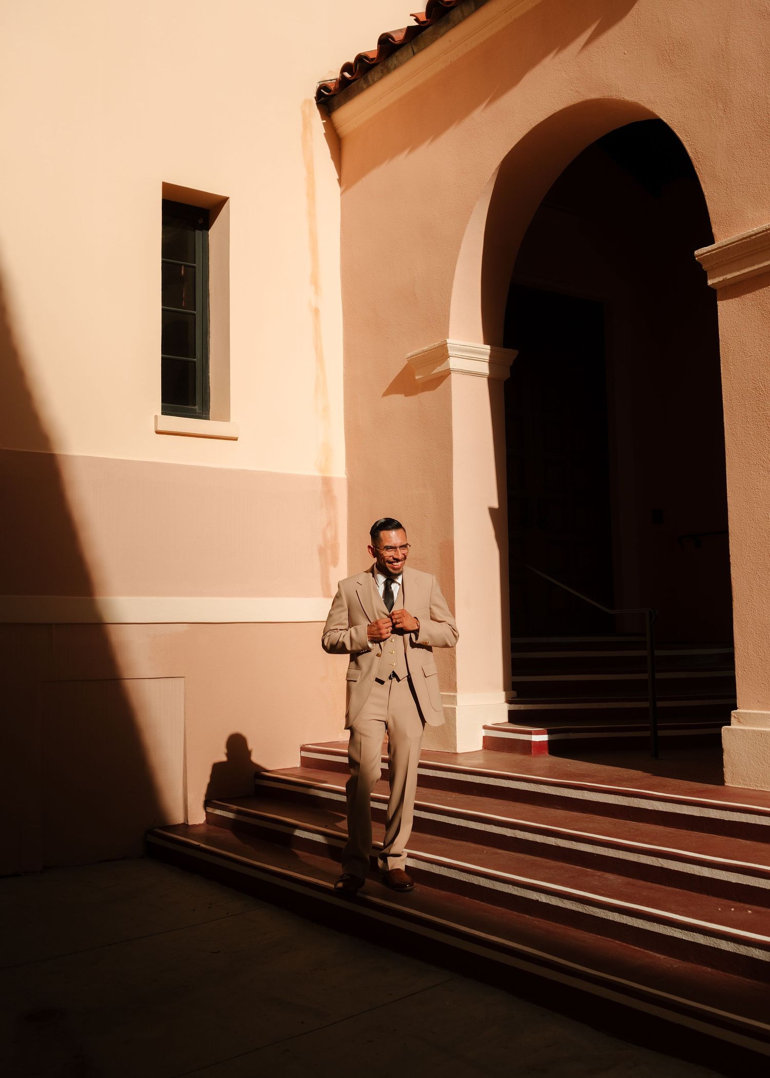 Man in beige suit standing on steps outside a pink arched building in warm golden light.
