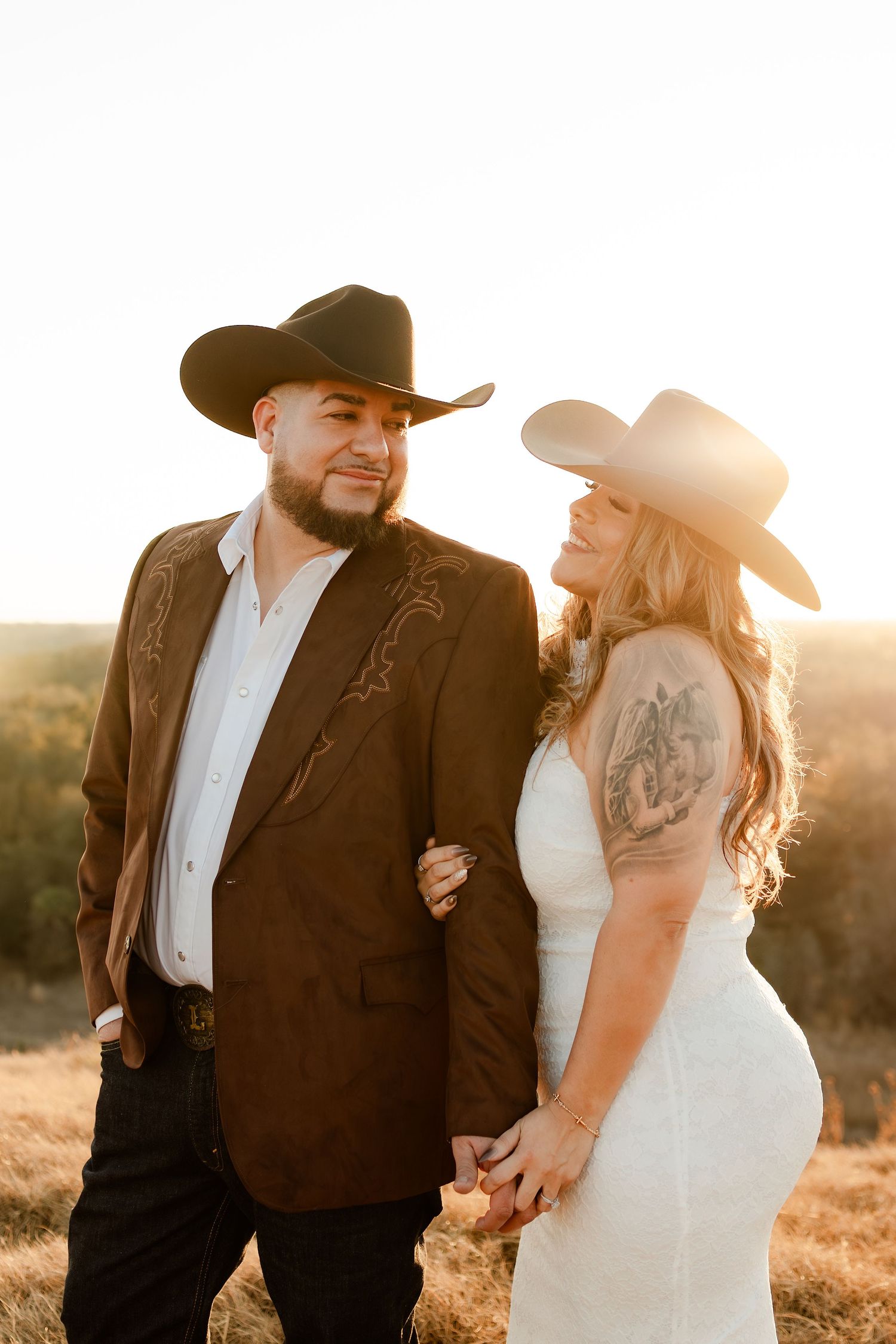 Couple in cowboy hats holding hands at golden sunset, man in brown western jacket, woman in white dress.