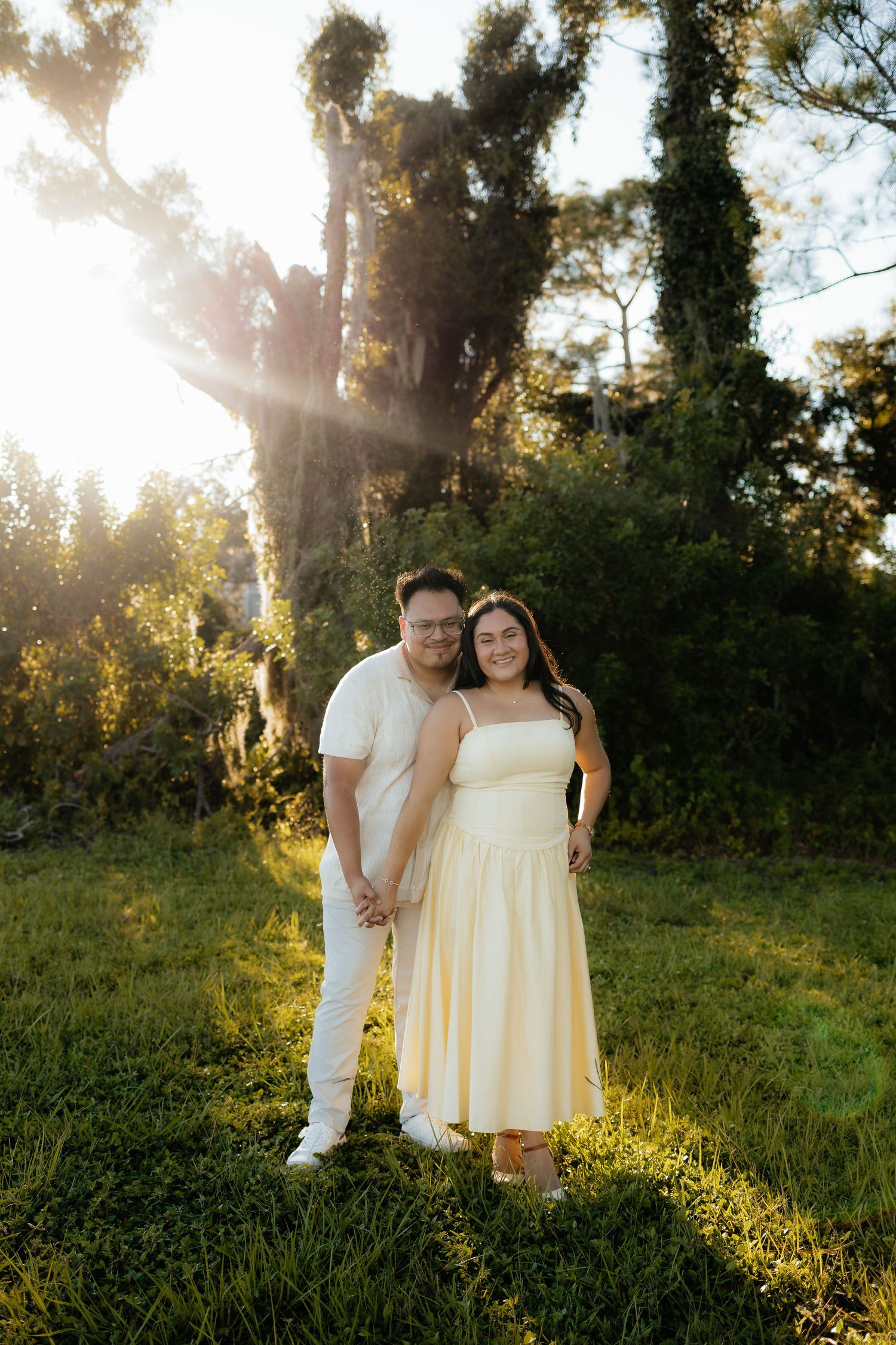 Couple holding hands in golden hour sunlight, woman in yellow dress and man in white outfit, standing in lush green park.