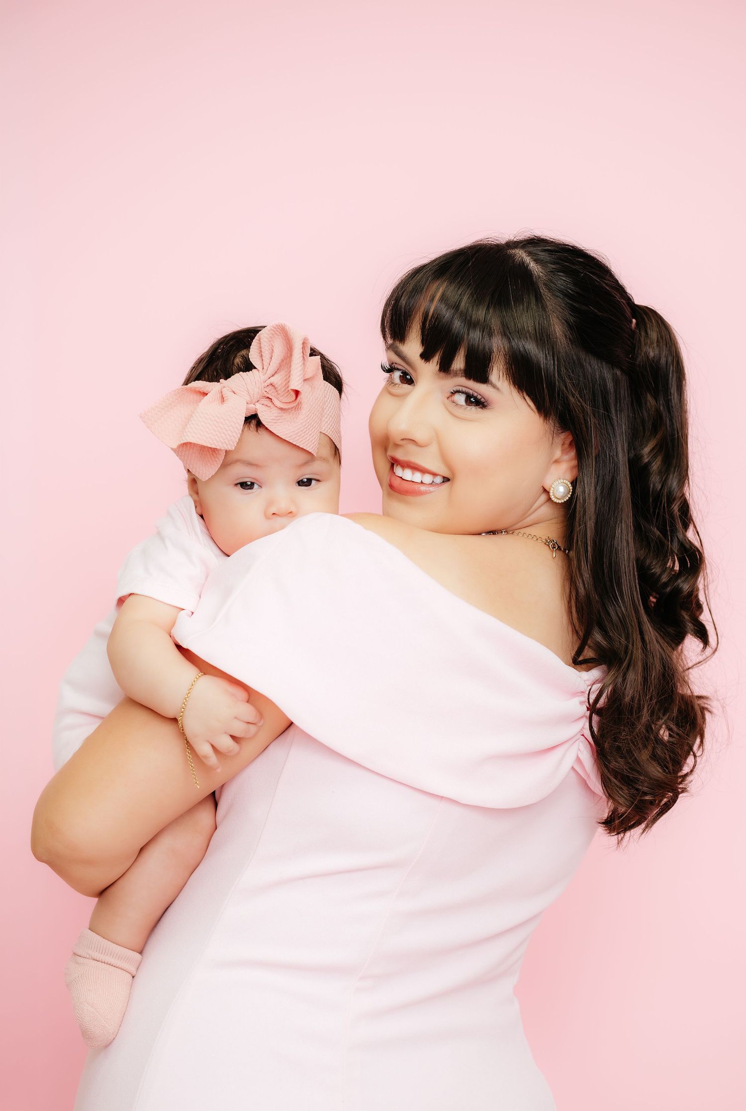 A smiling dark-haired woman in a pink off-shoulder top holds a baby girl with a pink bow on a pink background.