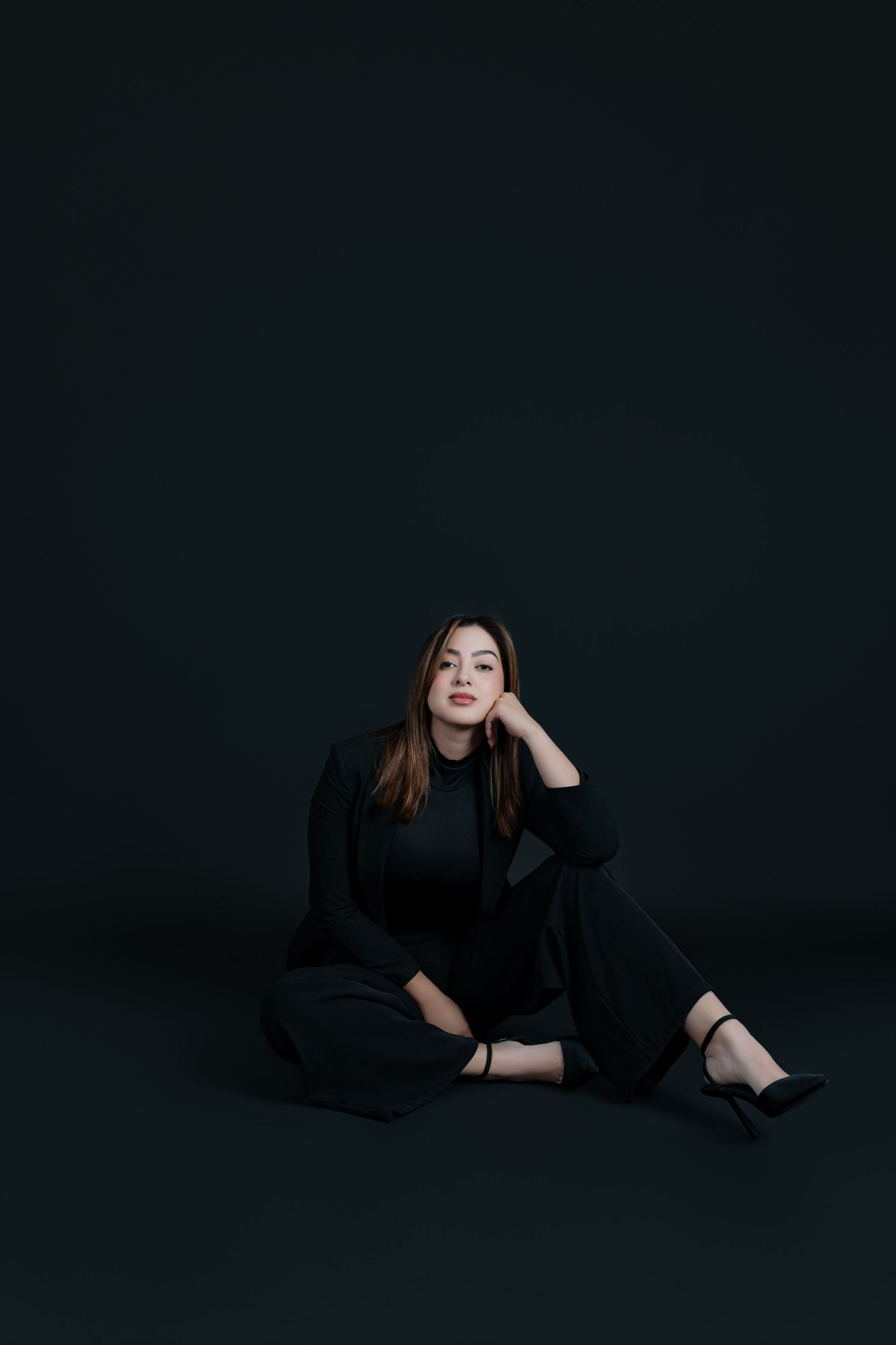Woman in all-black outfit sitting gracefully on floor against dark background, looking upward thoughtfully.