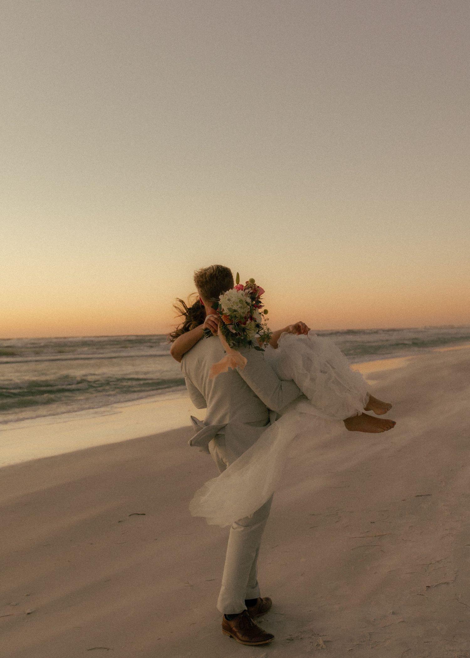 Groom lifts bride on sunset beach, she holds floral bouquet in white dress during romantic wedding photo.