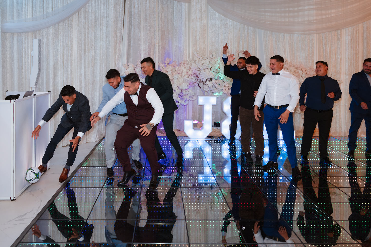 Groomsmen dancing and celebrating on a mirrored dance floor near illuminated J&K letters.