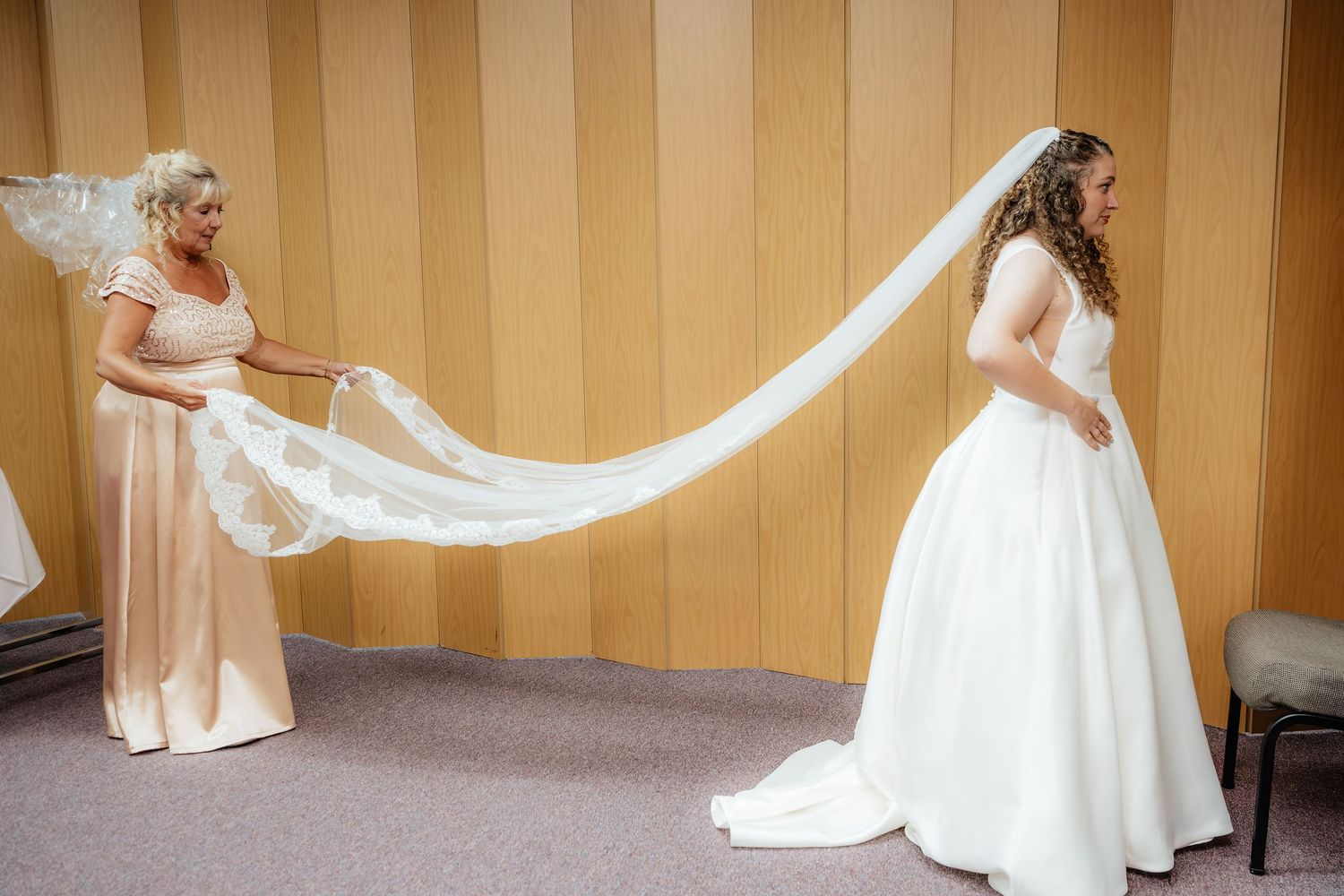 Bride in white gown with bridesmaid in pink dress holding long lace wedding veil in elegant wood-paneled room.