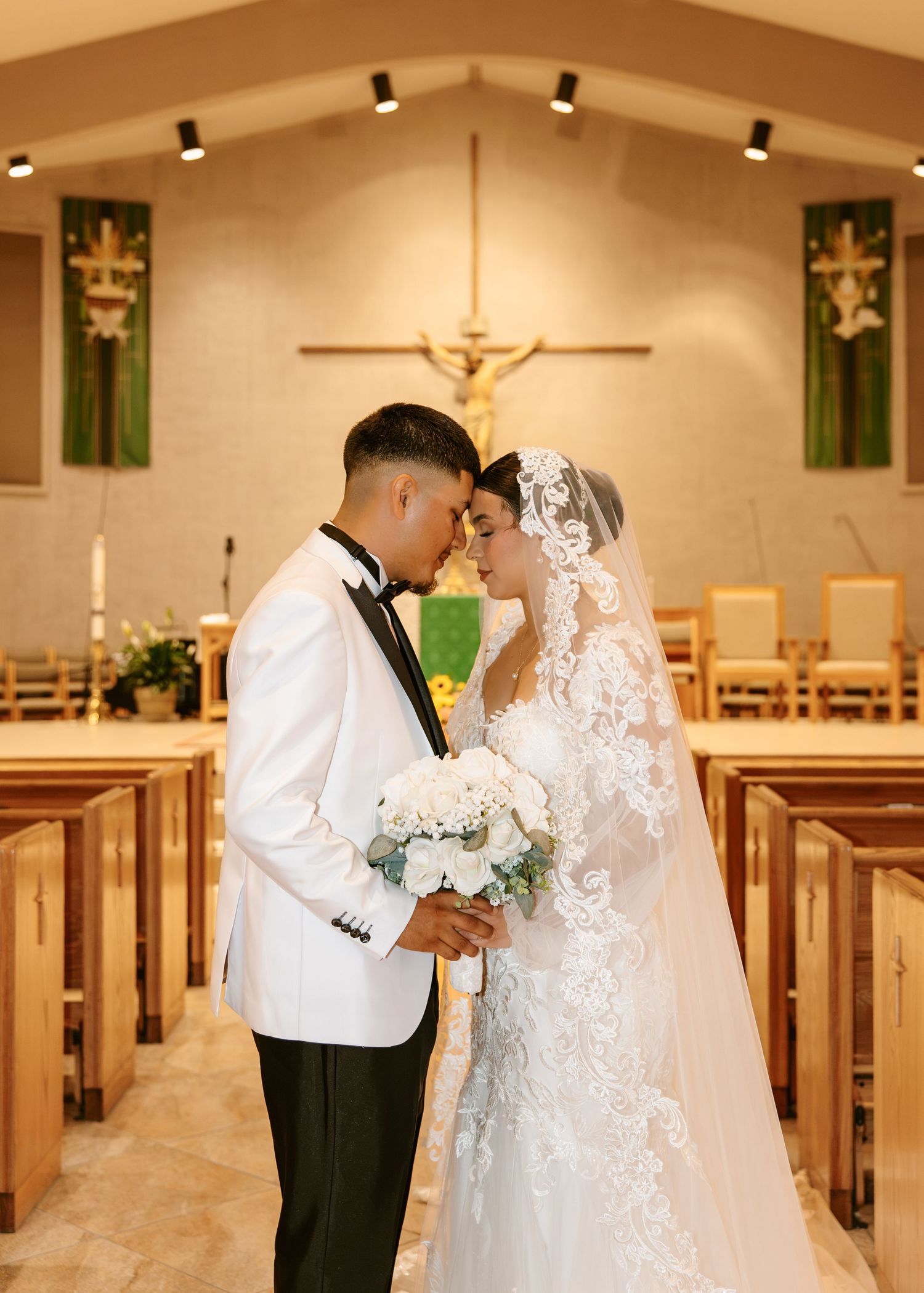 Bride and groom share an intimate moment in a Catholic church aisle, bride in lace gown with veil, groom in white tuxedo.