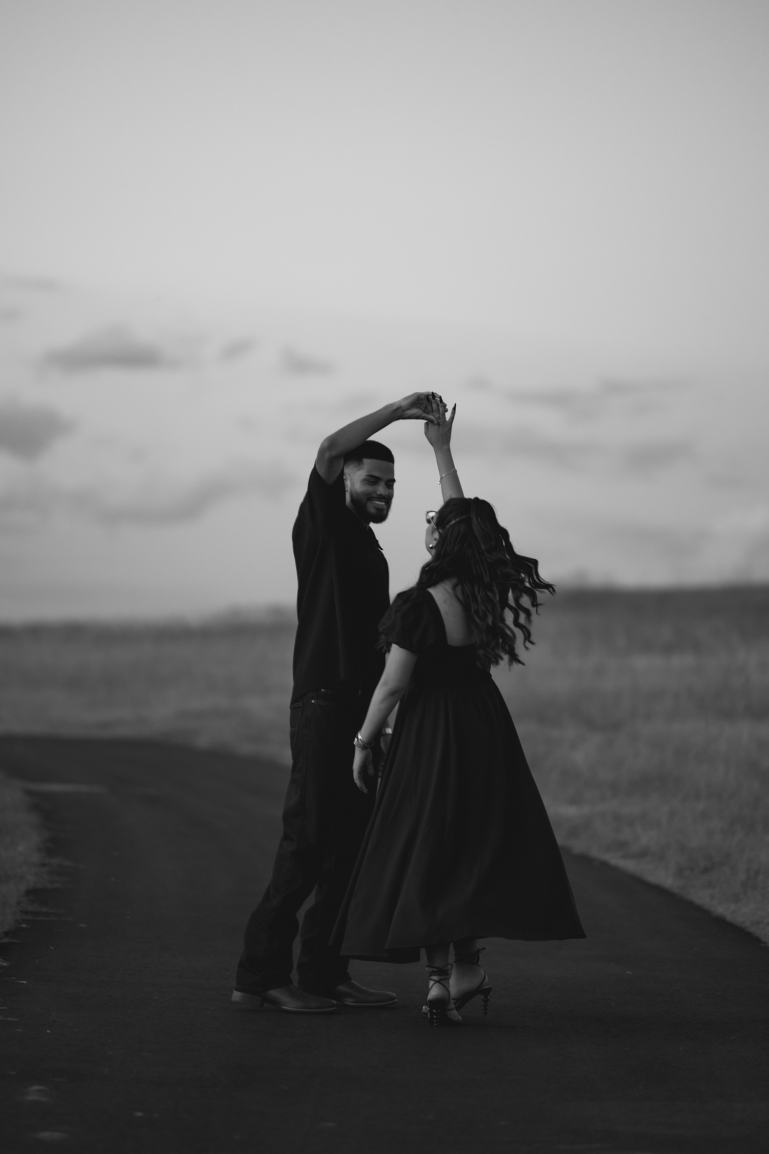 A romantic black and white silhouette of a couple dancing on a path against a cloudy sky.