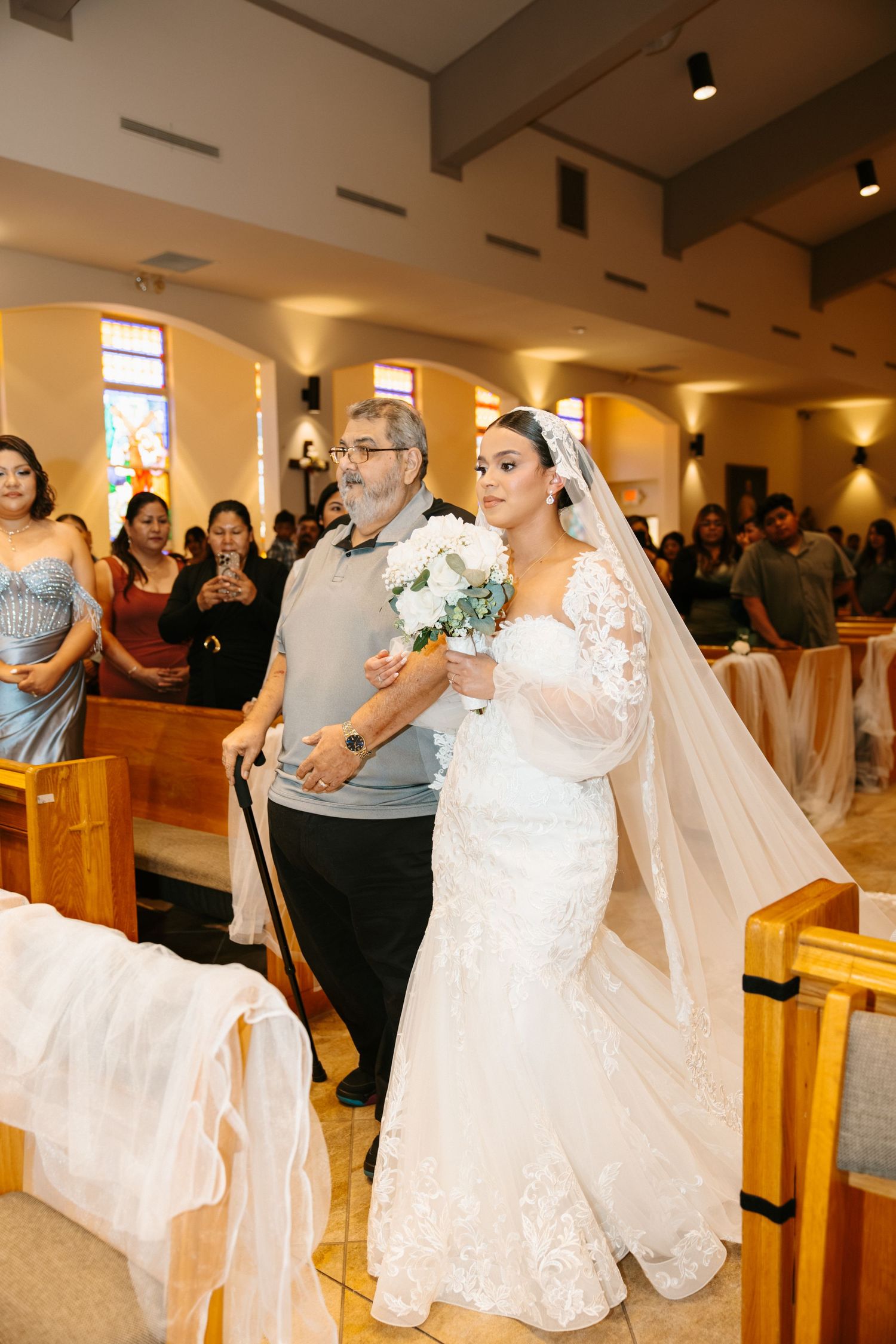 Bride in lace gown walks down church aisle with father using a cane, holding white bouquet.