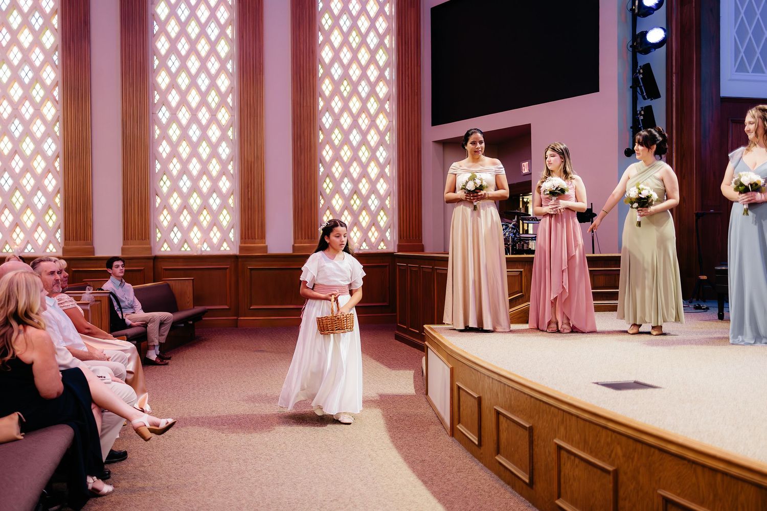 Flower girl in white dress walks down aisle as bridesmaids in pastel gowns stand on church stage.