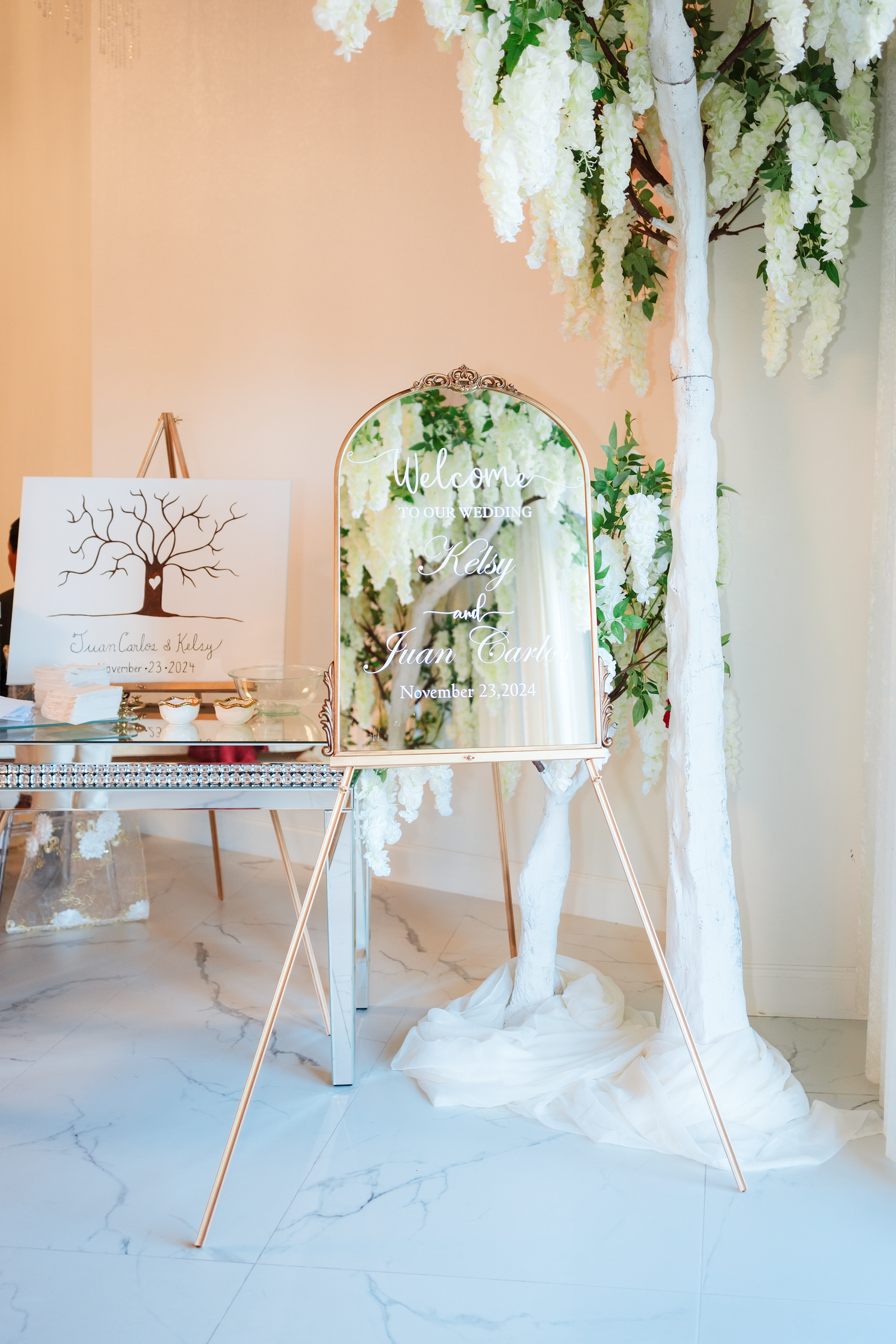 Elegant wedding welcome sign on gold easel with white floral arch and guest book display in soft pink venue.